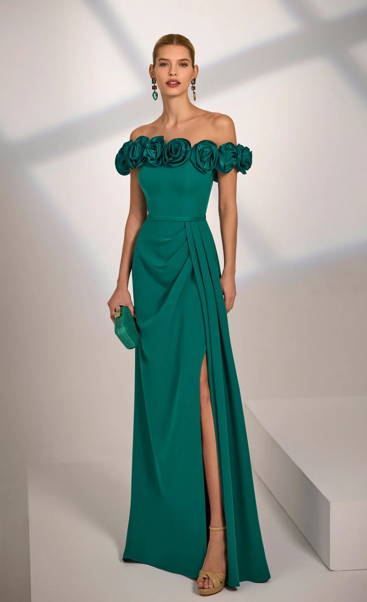 A woman wearing an off-shoulder green evening gown with large fabric roses around the neckline, a high slit, and beige heels, holding a small matching green clutch, against a neutral background.