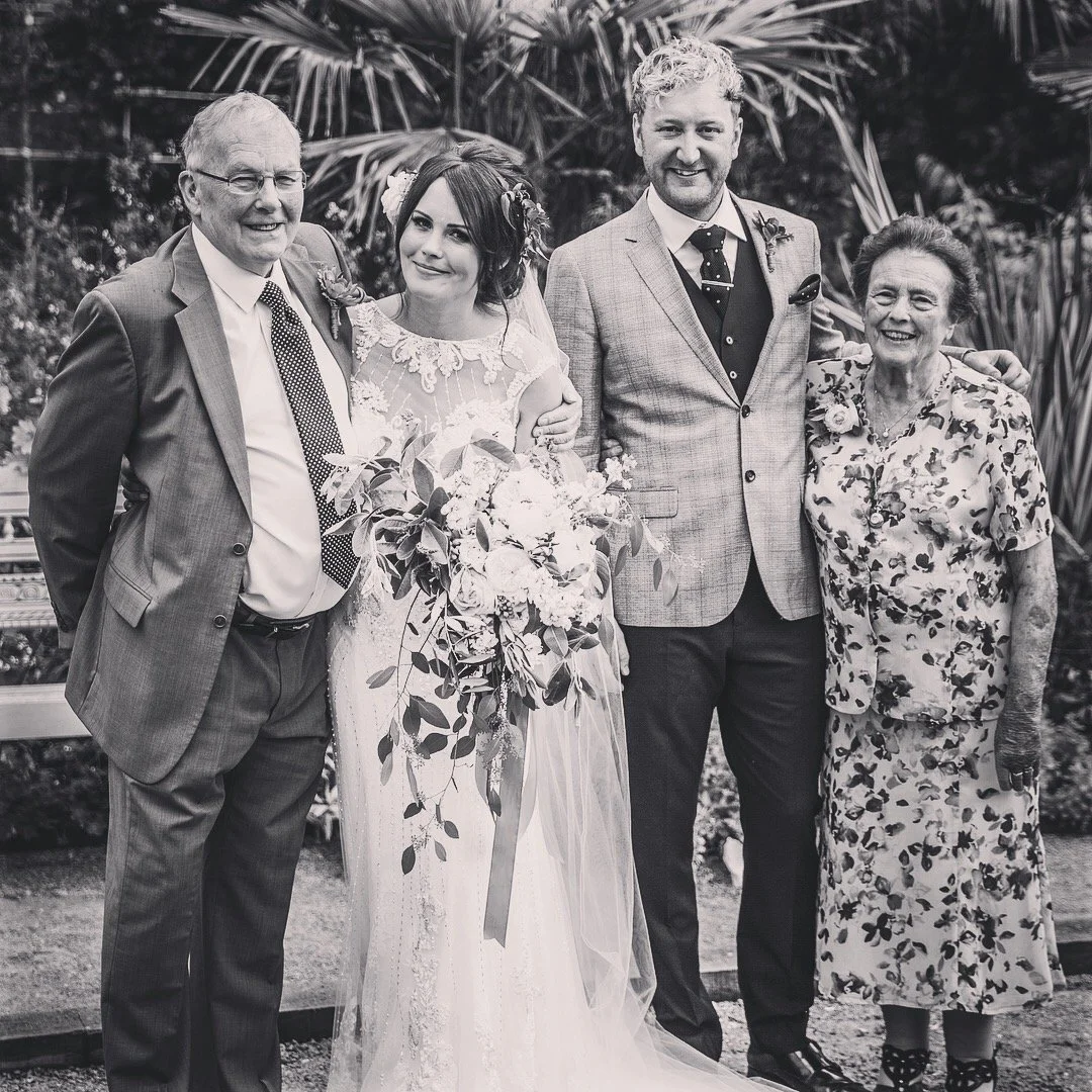 Black and white wedding photo of a bride in a lace dress holding a bouquet, standing with three men and an older woman, outdoors with palm leaves in the background.