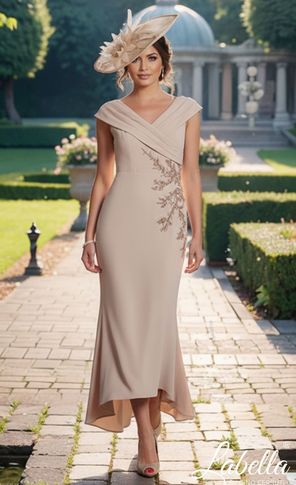 A woman in an elegant beige evening gown with intricate embroidery, accessorized with a large matching hat, walking outdoors on a stone pathway in a garden setting.