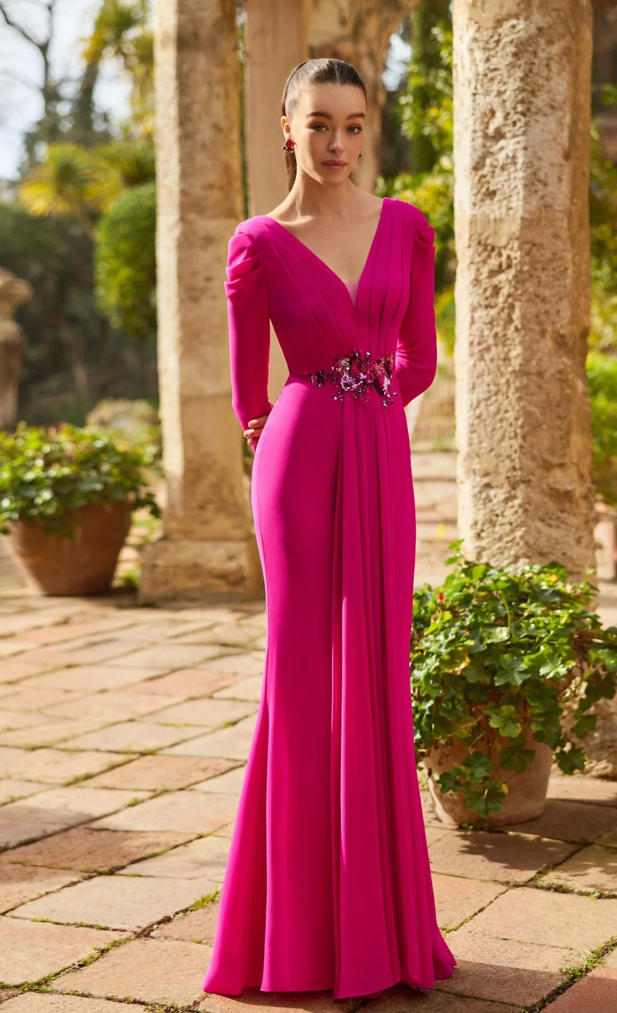 A woman in a long, vibrant pink gown with detailed embroidery at the waist, standing outdoors among stone columns and greenery.
