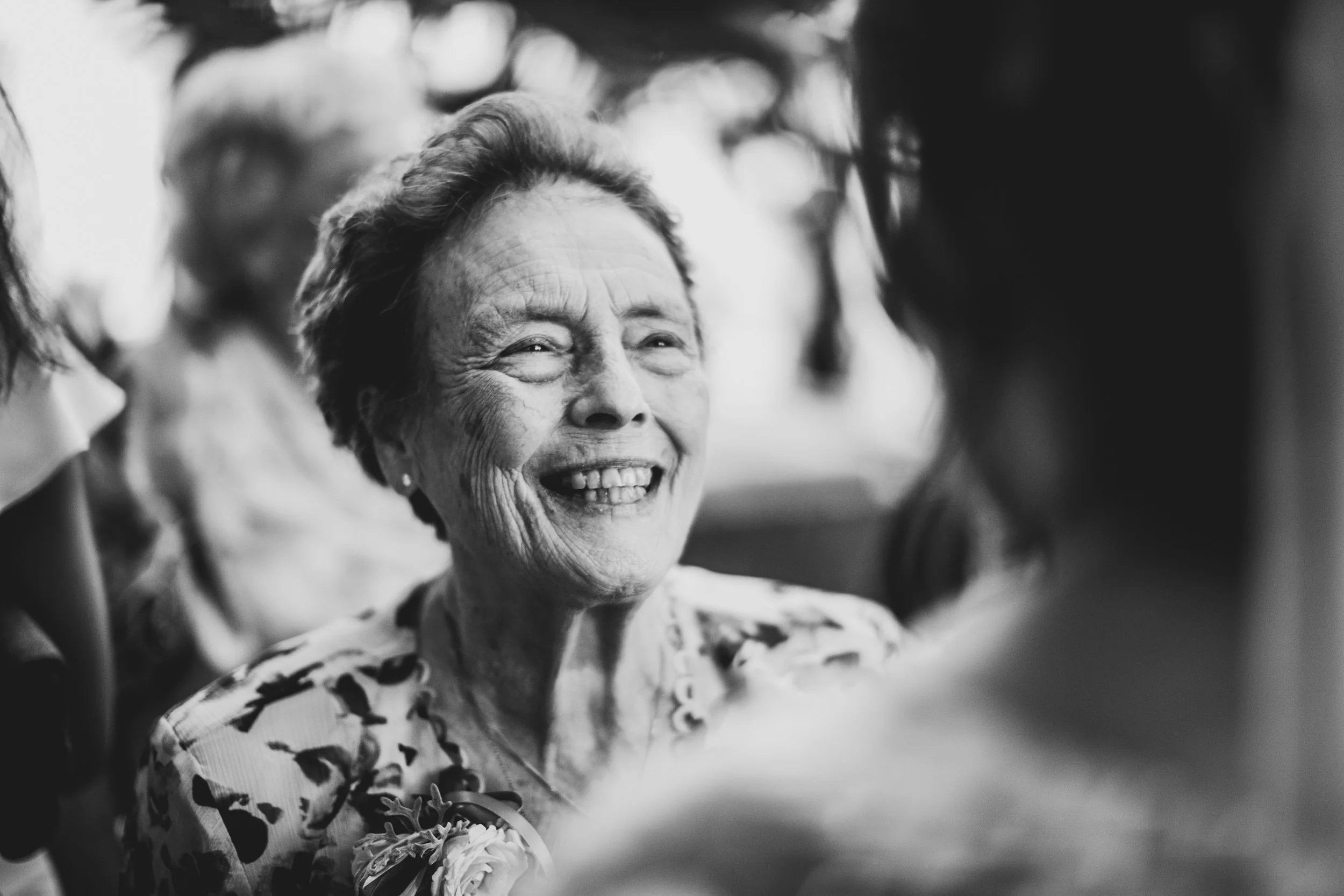 An elderly woman smiling, dressed in a floral patterned top, engaging happily with others around her, in a candid black and white photograph.