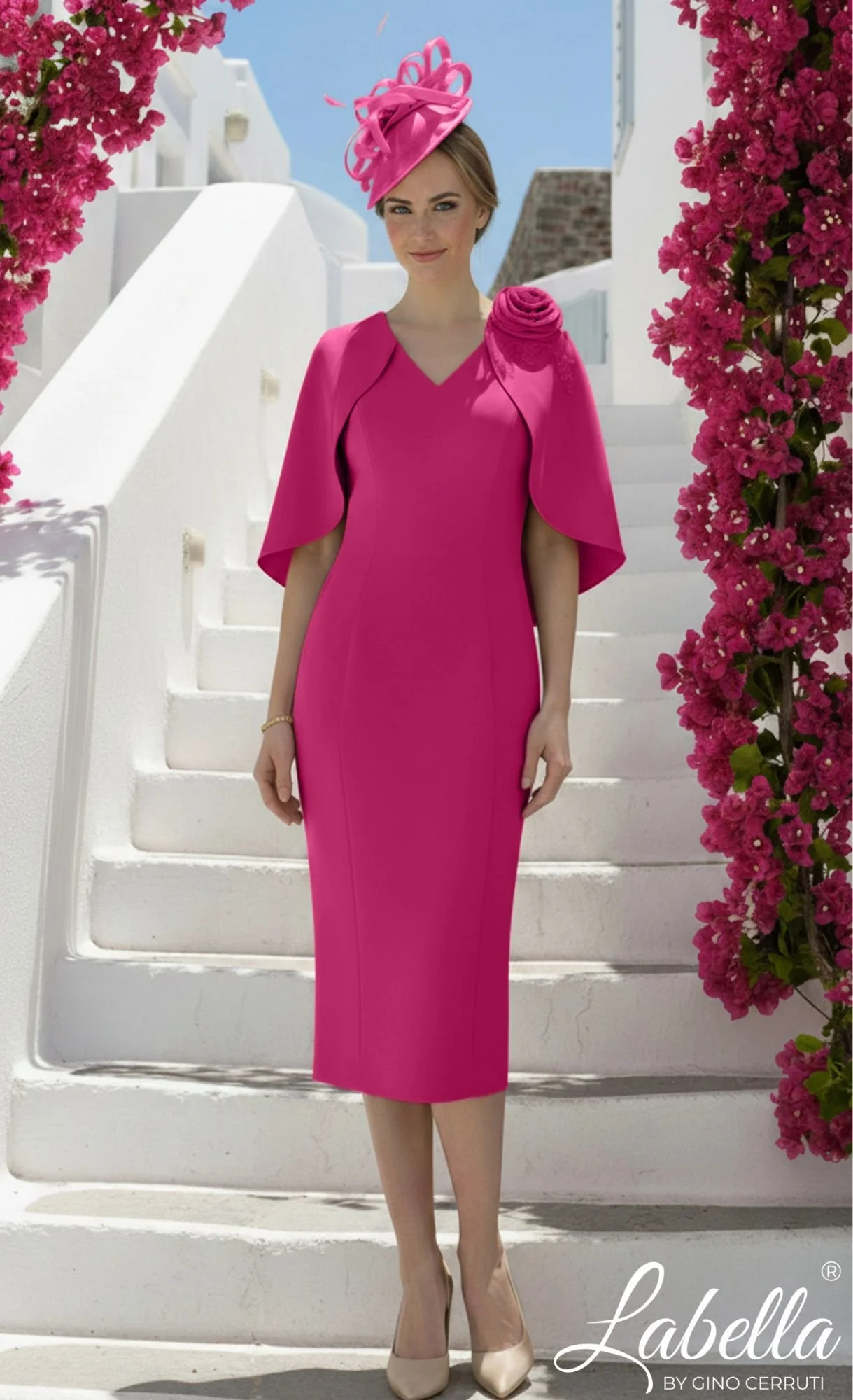 A woman wearing a bright pink dress and matching hat stands on white stairs in front of pink flowering bushes under a blue sky.
