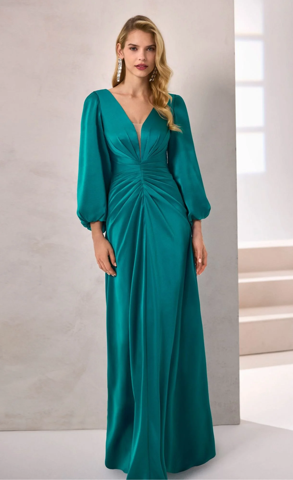 A woman in a teal, long, elegant dress with puffed sleeves standing indoors with a neutral background and stairs.