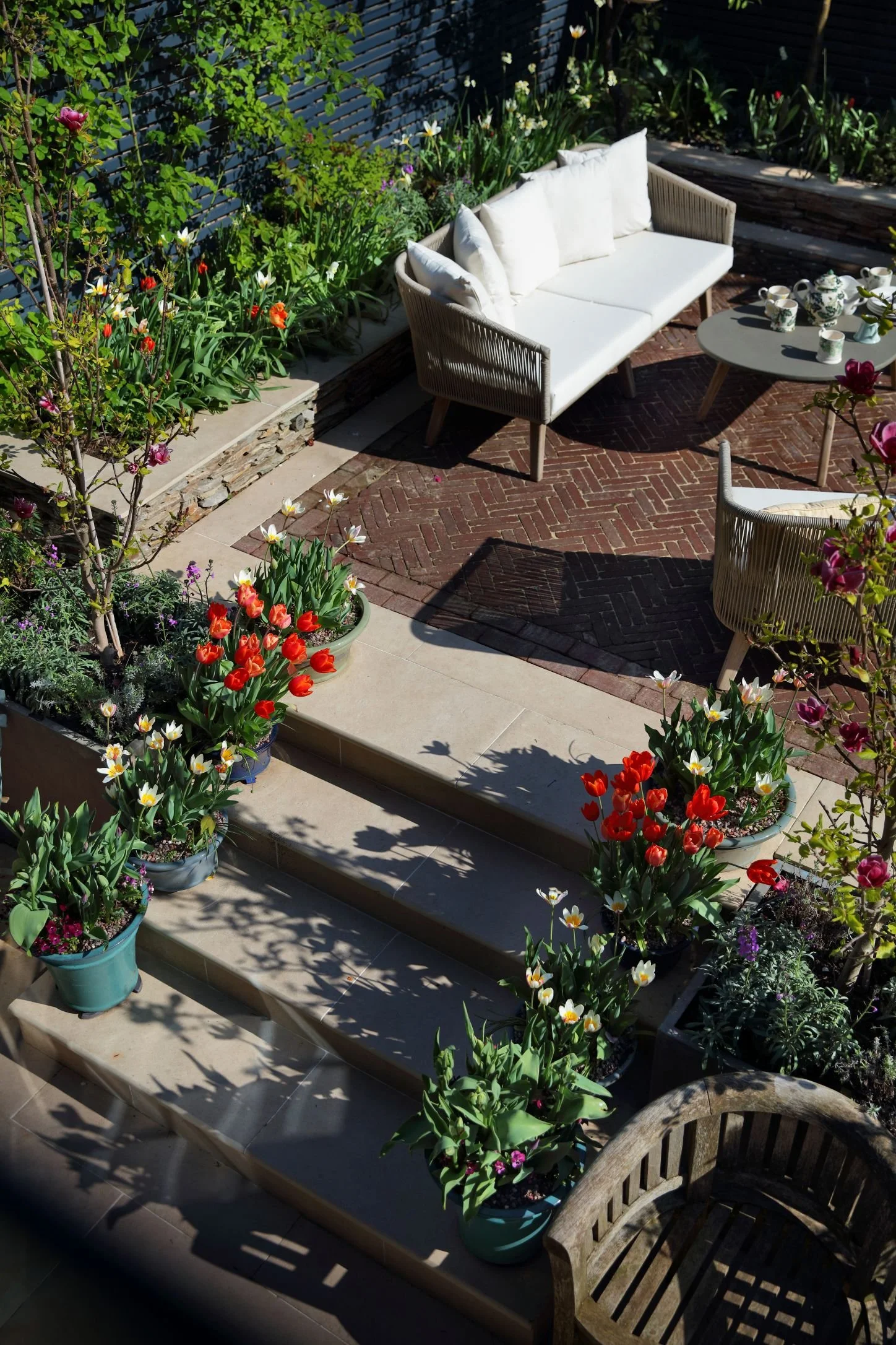 A cozy outdoor patio with a white cushioned sofa, a round table with a tea set, and colorful potted flowers on steps leading up to the seating area, surrounded by lush greenery and flowers.