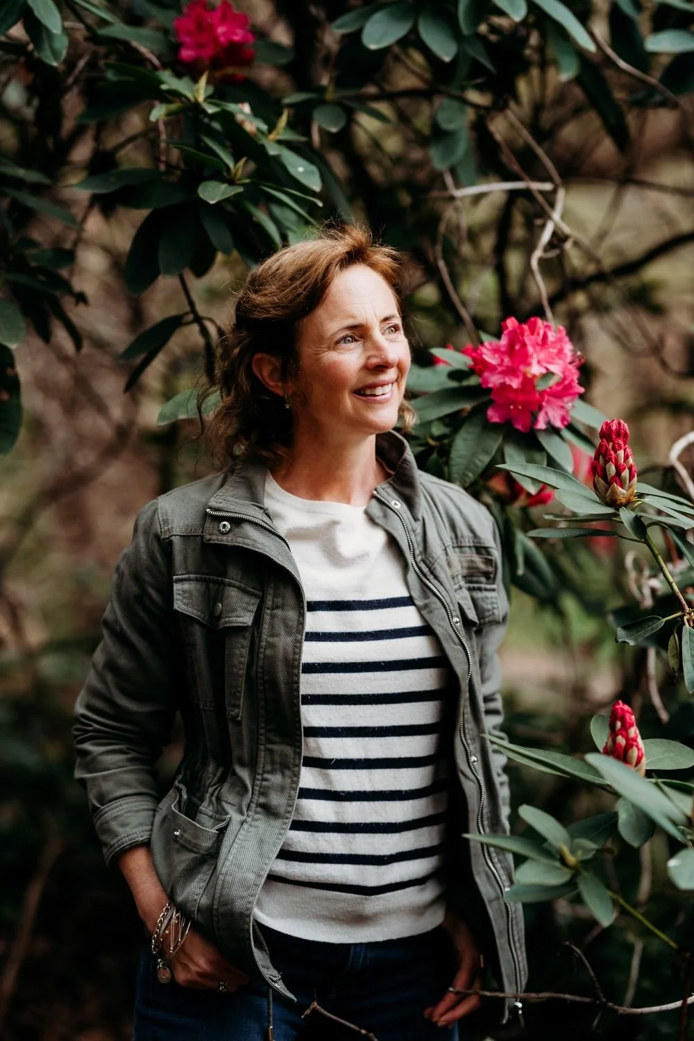 A woman with short brown hair smiling and looking to the side, standing outdoors among green leaves and pink rhododendron flowers.