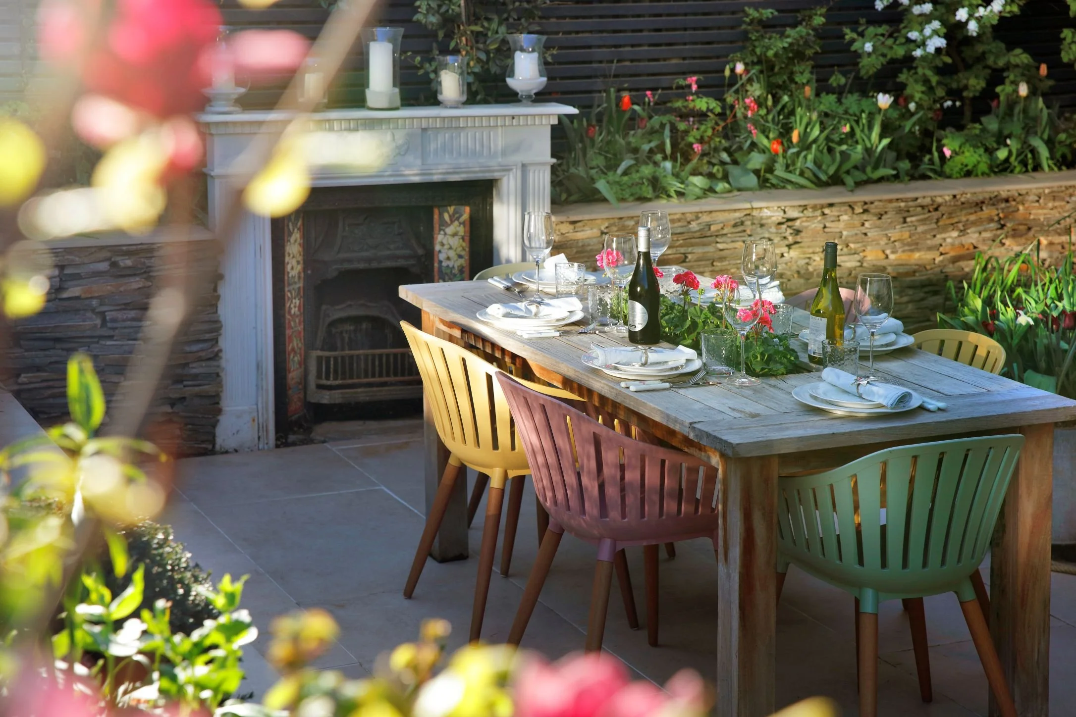 A cozy backyard garden with blooming tulips and other flowers, a white outdoor sofa with cushions, and a high fence with trees and flowering plants in the background.