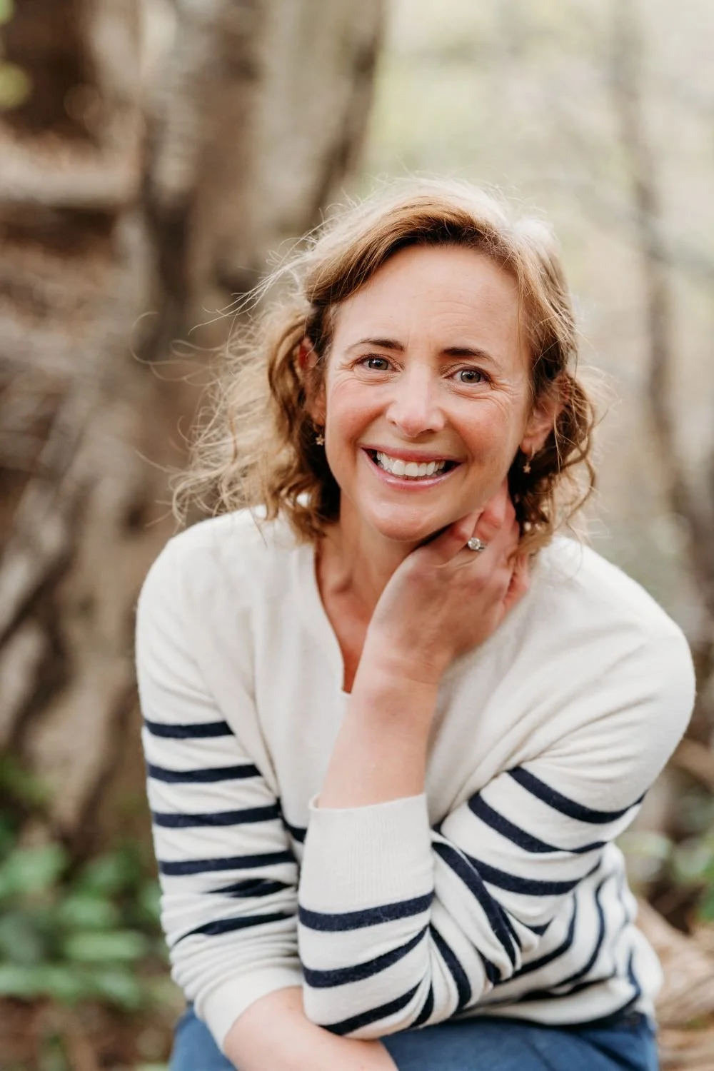 A woman with curly, shoulder-length hair smiling outdoors with trees and blurred background, wearing a white and navy striped sweater.
