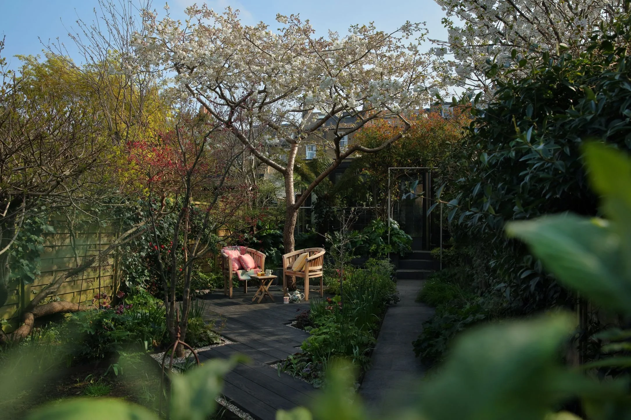 A garden with colorful flowers in focus at the front and a lawn with a black wooden fence in the background.