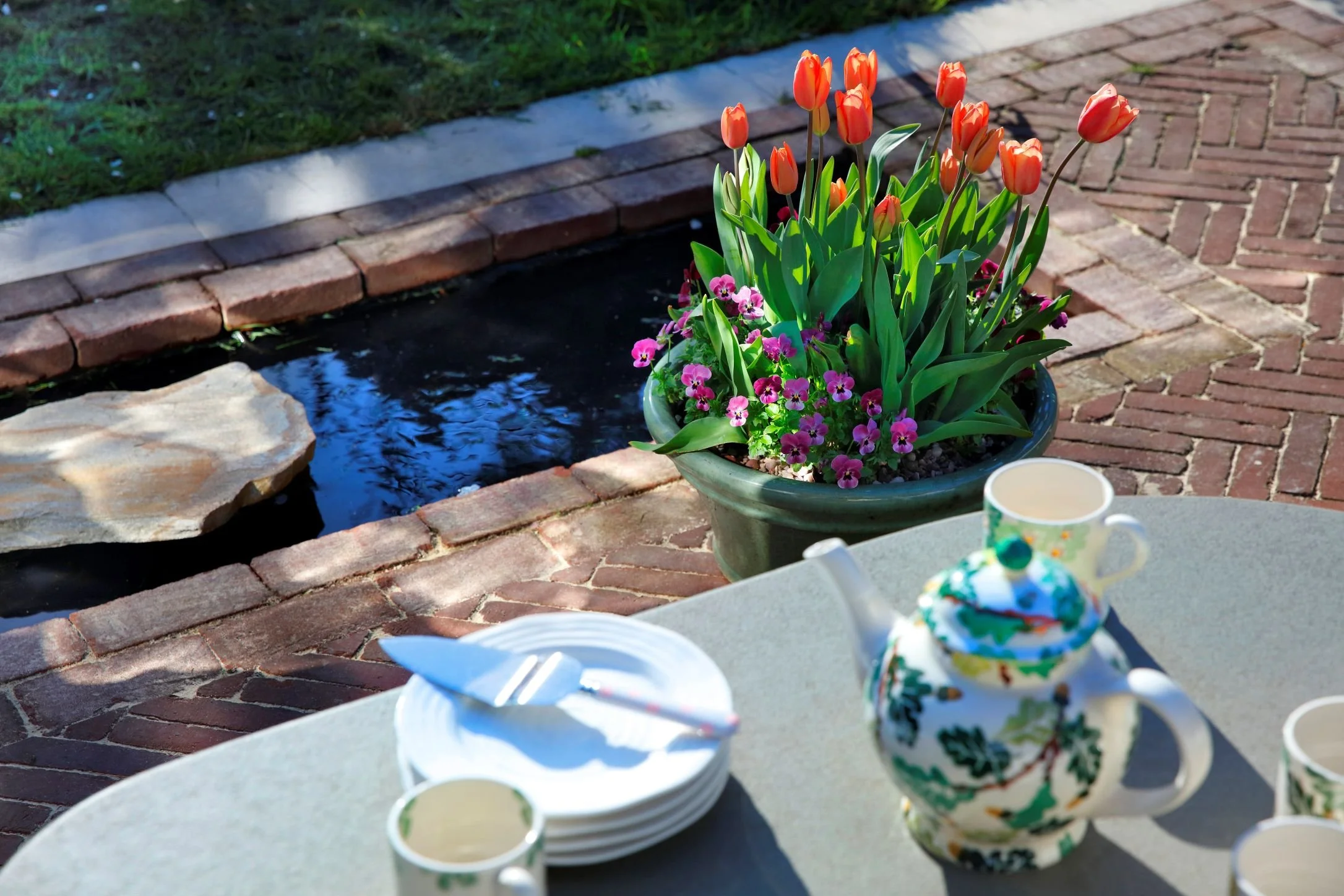 A flower pot with orange tulips and pink and purple small flowers on a patio next to a small pond, with a white marble border, and a table set with cups, plates, and a colorful teapot in the foreground.