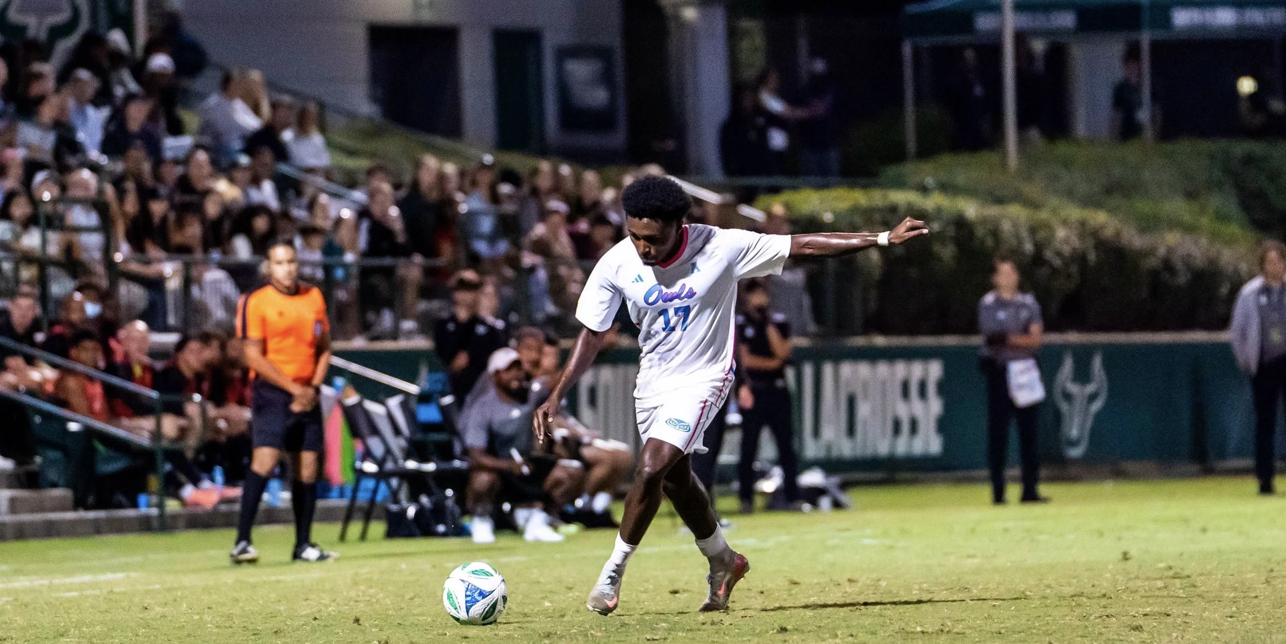 A soccer player wearing a white uniform with the number 17 is about to kick a soccer ball on a field, with a crowd of spectators watching from the stands during a game.