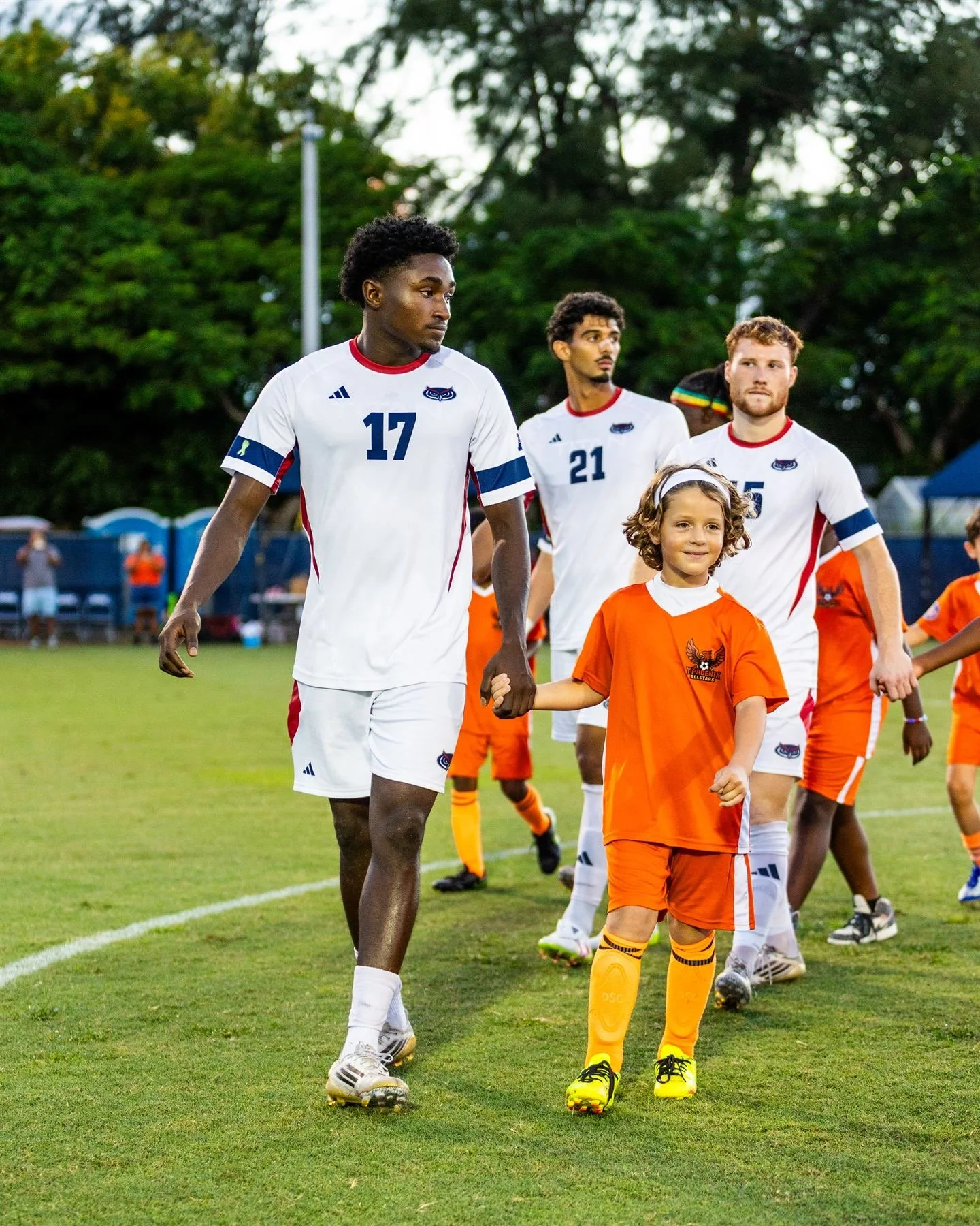 Soccer players in white uniforms walking with a young girl in an orange sports jersey on a soccer field.