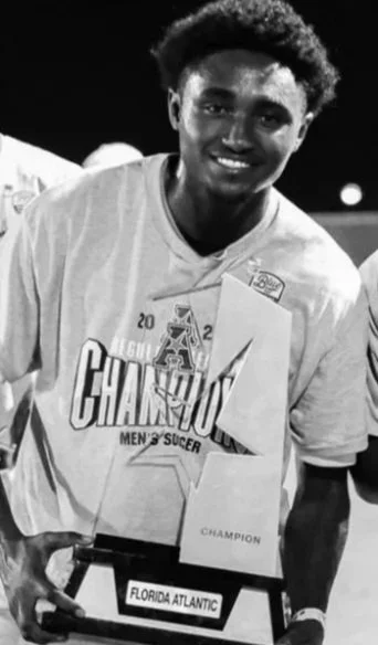Young man smiling, wearing a soccer jersey with a championship medal around his neck, holding a trophy that says 'Champion' and a plaque that reads 'Florida Atlantic'.