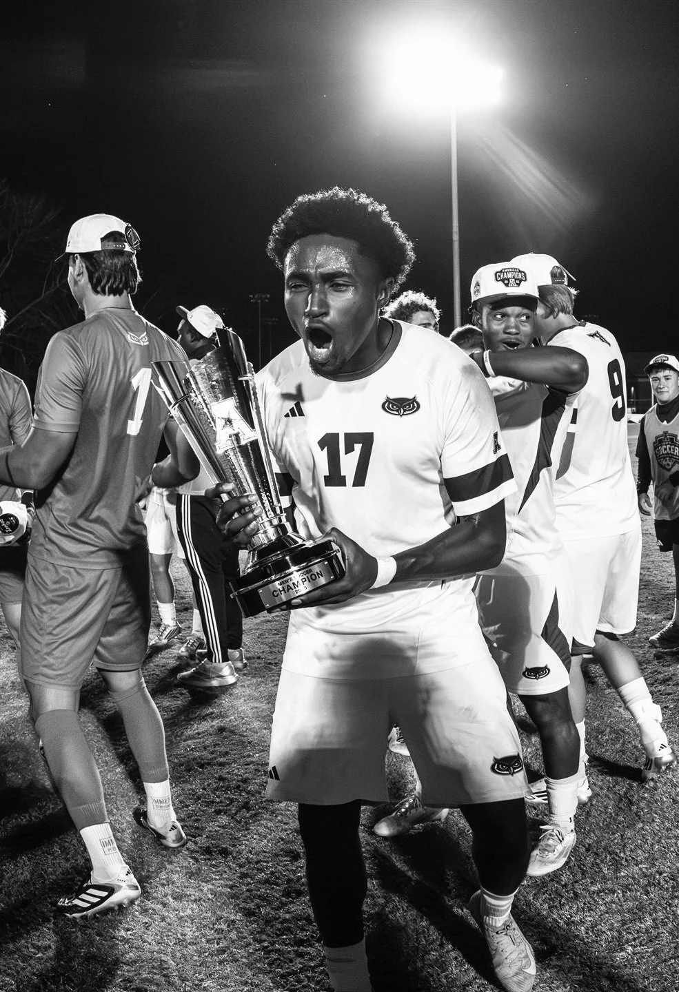 Black and white photo of a soccer player holding a trophy, celebrating victory with teammates at night on a soccer field, stadium lights overhead.