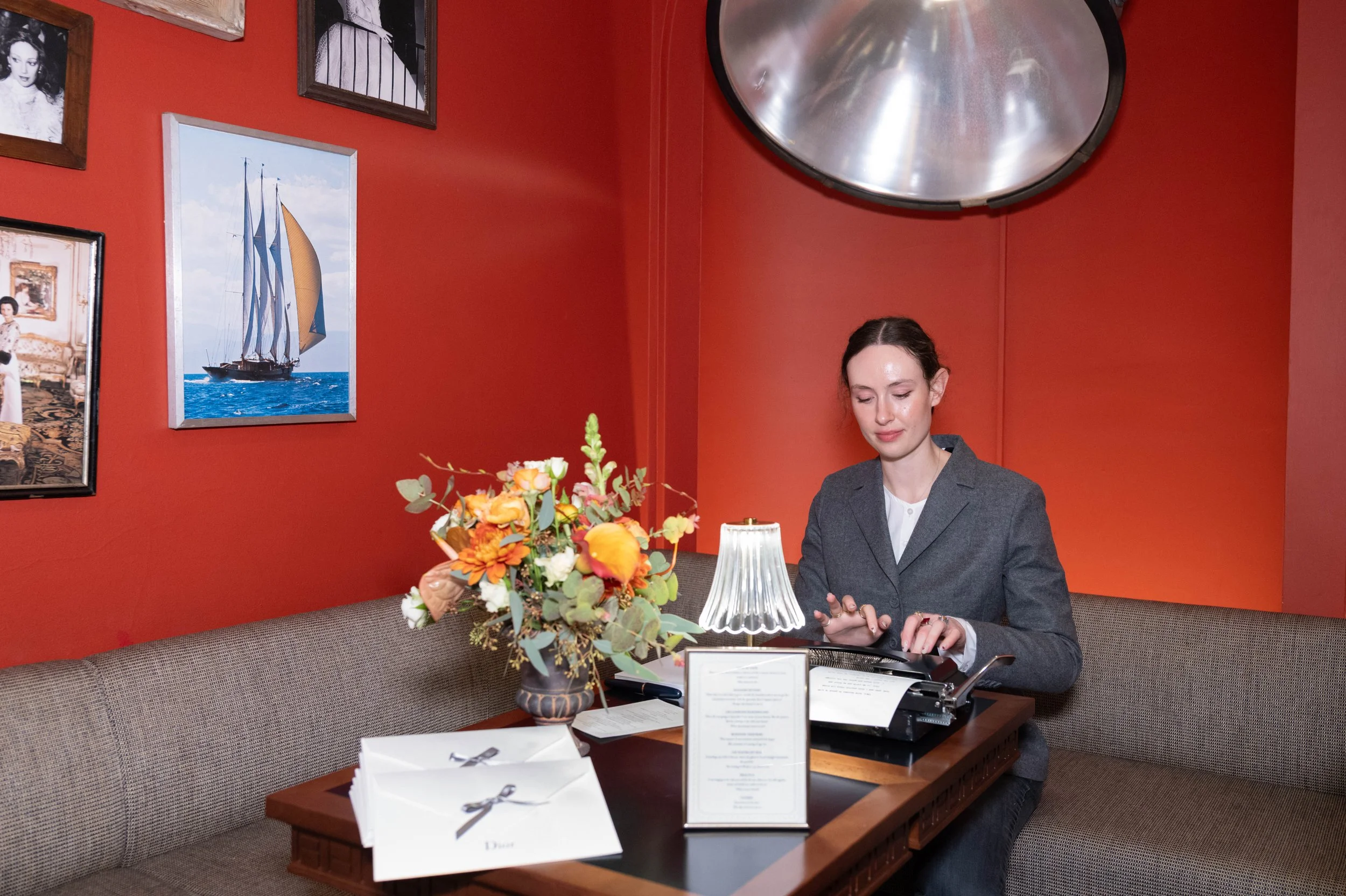 A woman in a gray blazer sitting at a booth in a restaurant, typing on a typewriter, with a floral centerpiece, a lamp, and menus on the table, red walls decorated with framed photos and artwork, and a large metallic light fixture overhead.