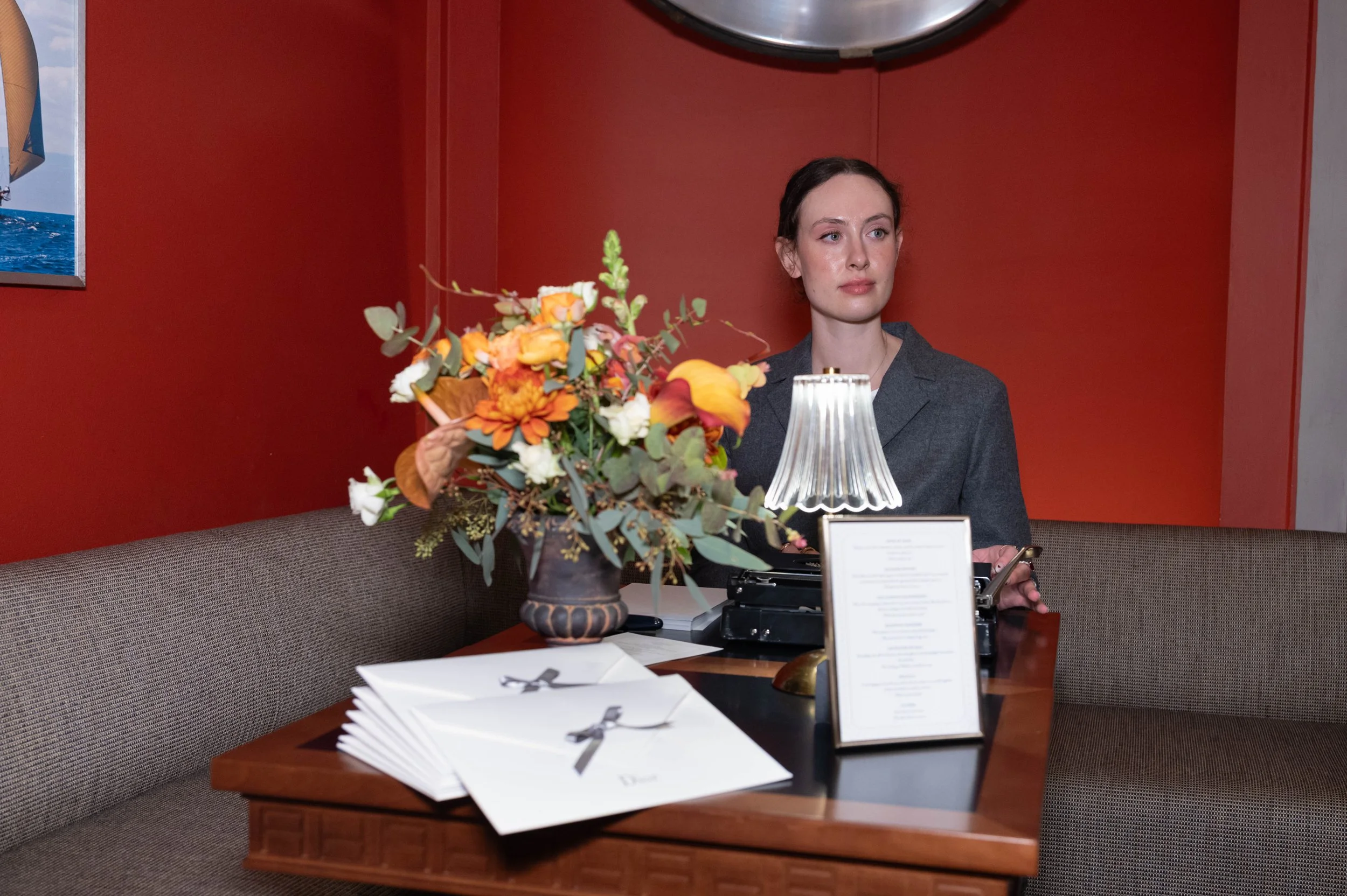 A woman with dark hair and light skin sitting at a table with a floral arrangement, lamp, menu, and a typewriter, against a red wall.