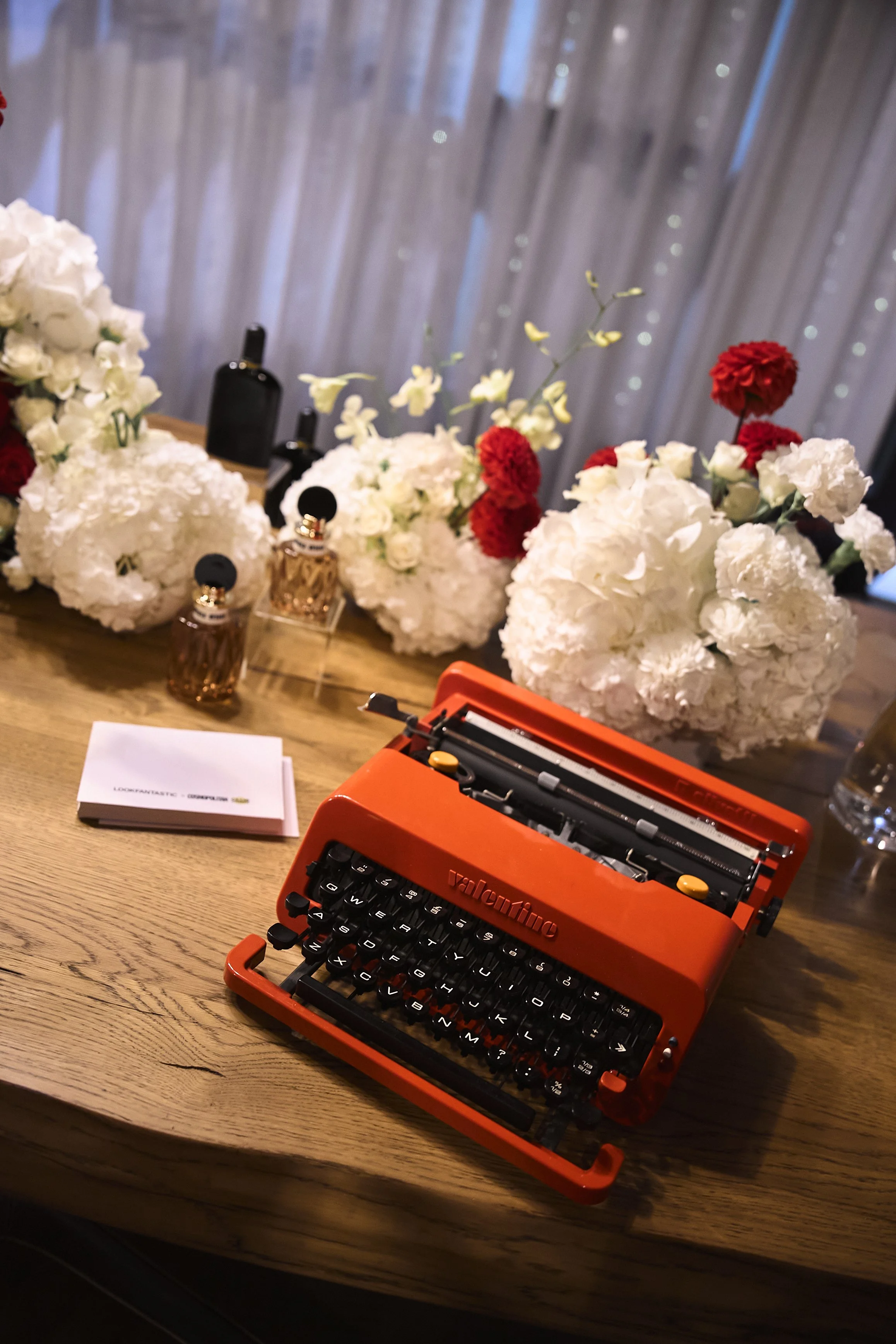 A vintage orange typwriter on a wooden table, surrounded by white and red flower bouquets, perfume bottles, a small notebook, and various bottles, with a sheer curtain in the background.
