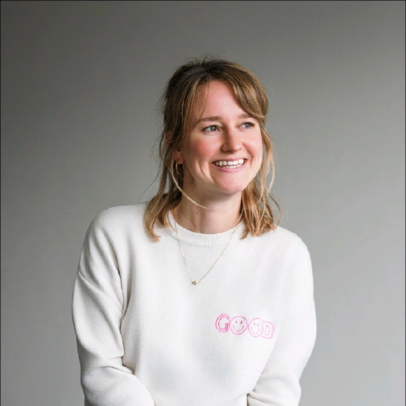 Een jonge vrouw lacht terwijl ze naar rechts kijkt, draagt een witte trui met het woord 'GOOD' en een smiley erop, en heeft oorbellen en een ketting.