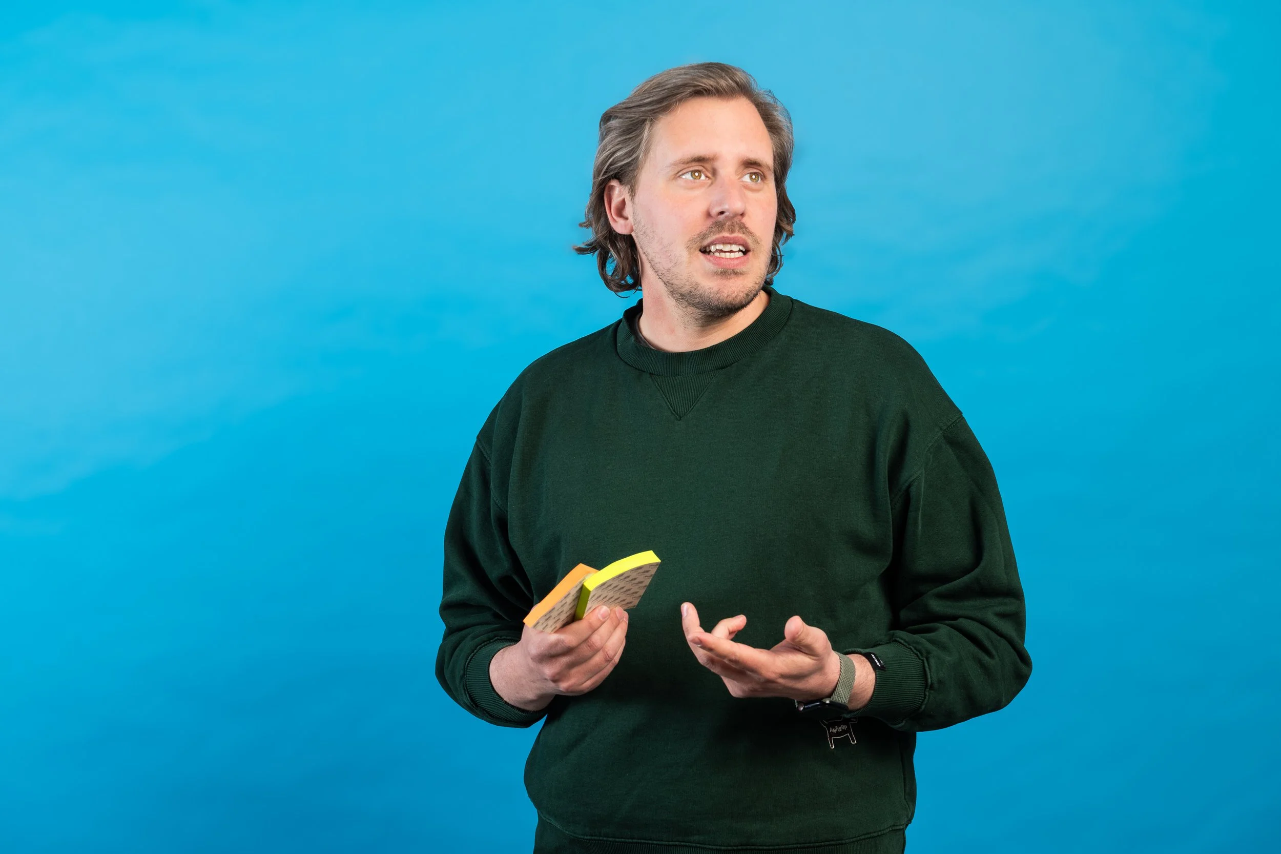 Man met lang bruin haar en een groen sweatshirt houdt een stapel speelkaarten vast, staand voor een blauw achtergrond.