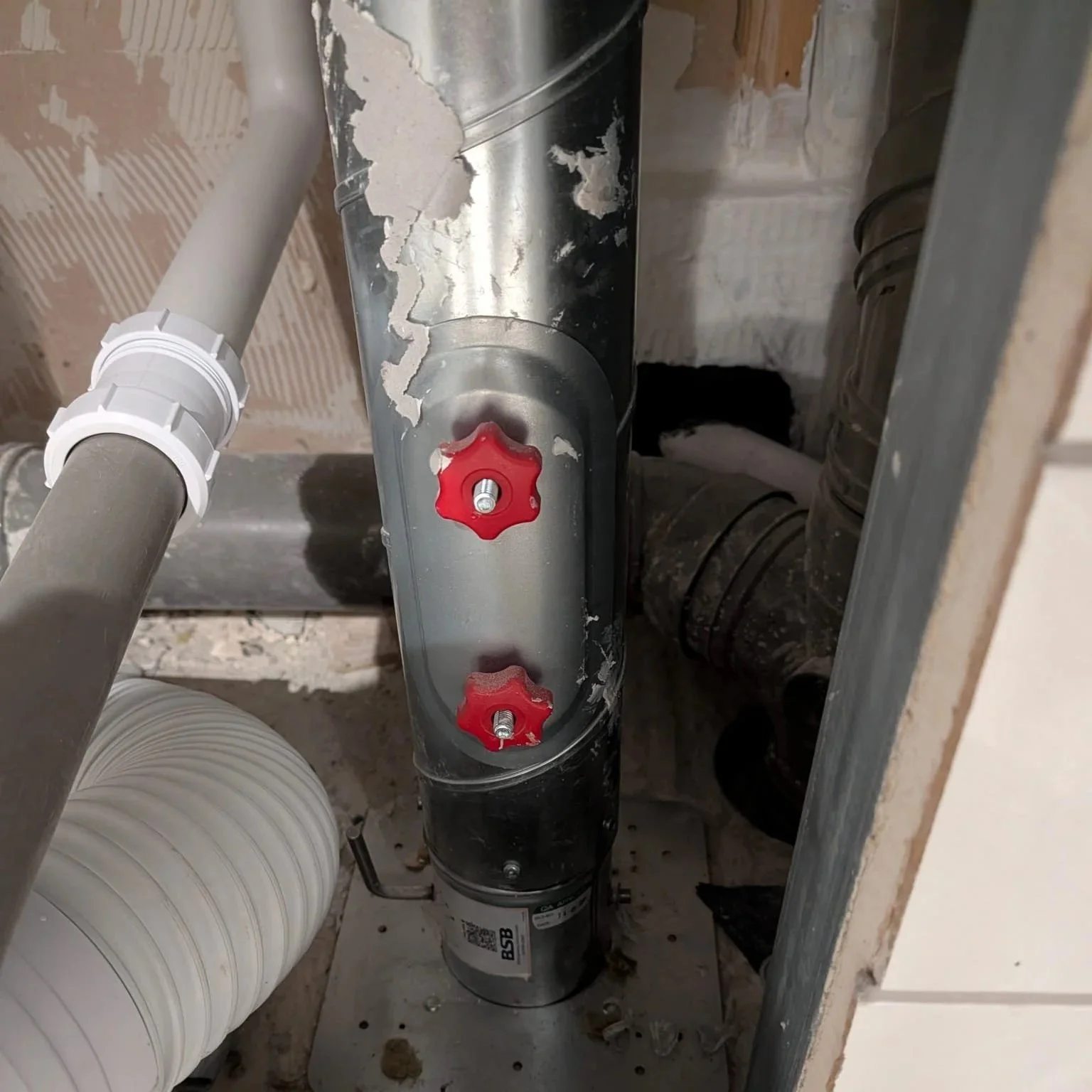 Close-up of a drainage pipe with two red valve handles, surrounded by metal and plastic pipes in a construction or repair setting.