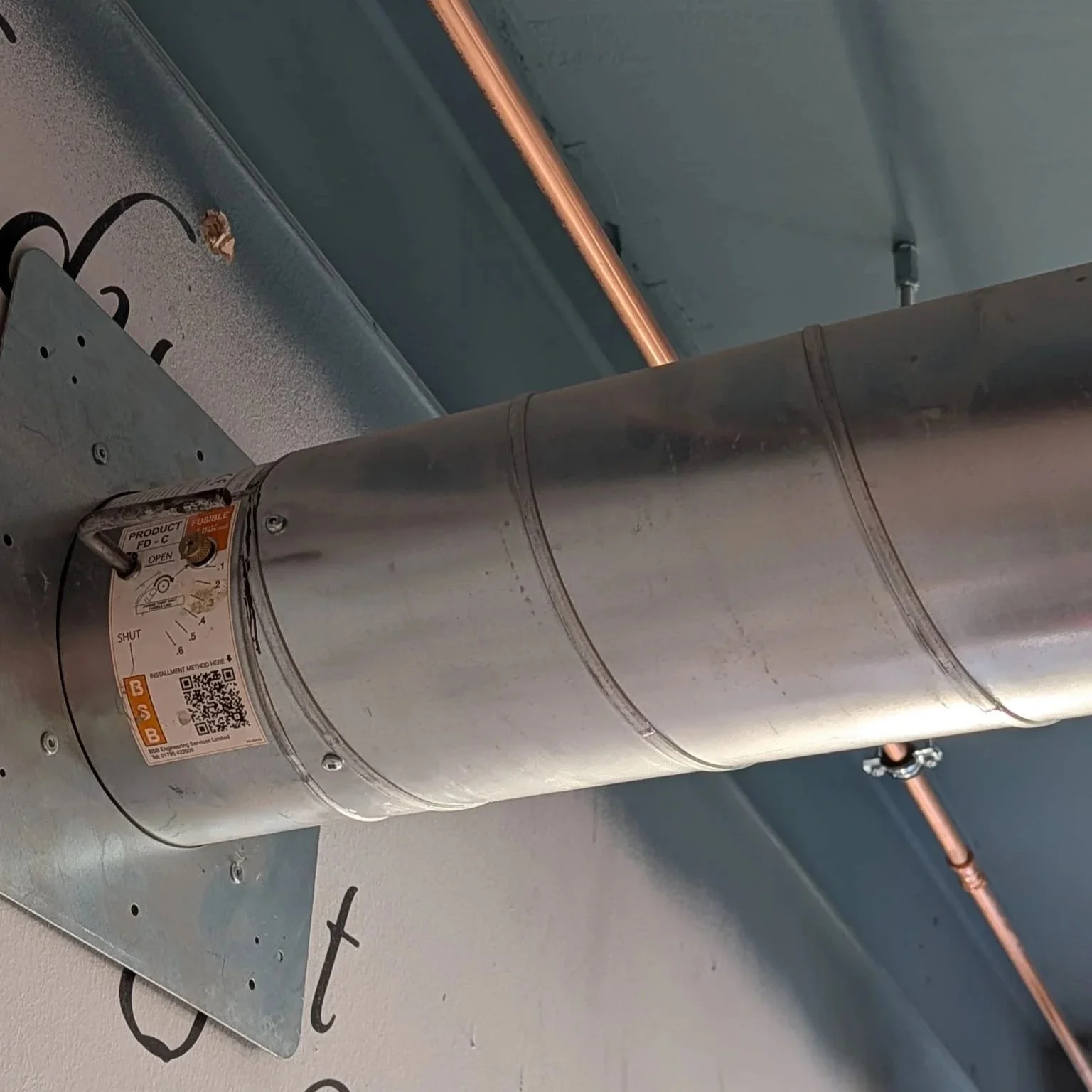 Close-up of an industrial metal duct with a QR code and warning label, mounted on a wall with a ceiling and copper pipe visible in the background.