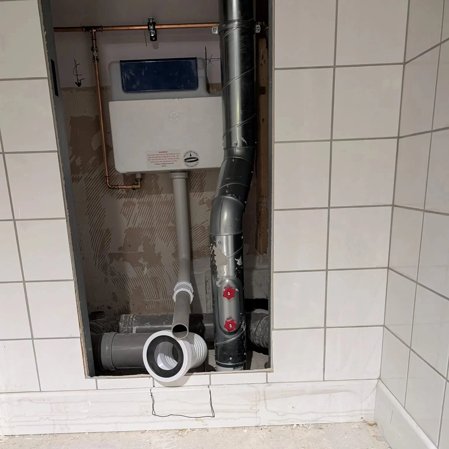 Open wall with plumbing and electrical components inside a small utility opening, including pipes, valves, and a water heater.