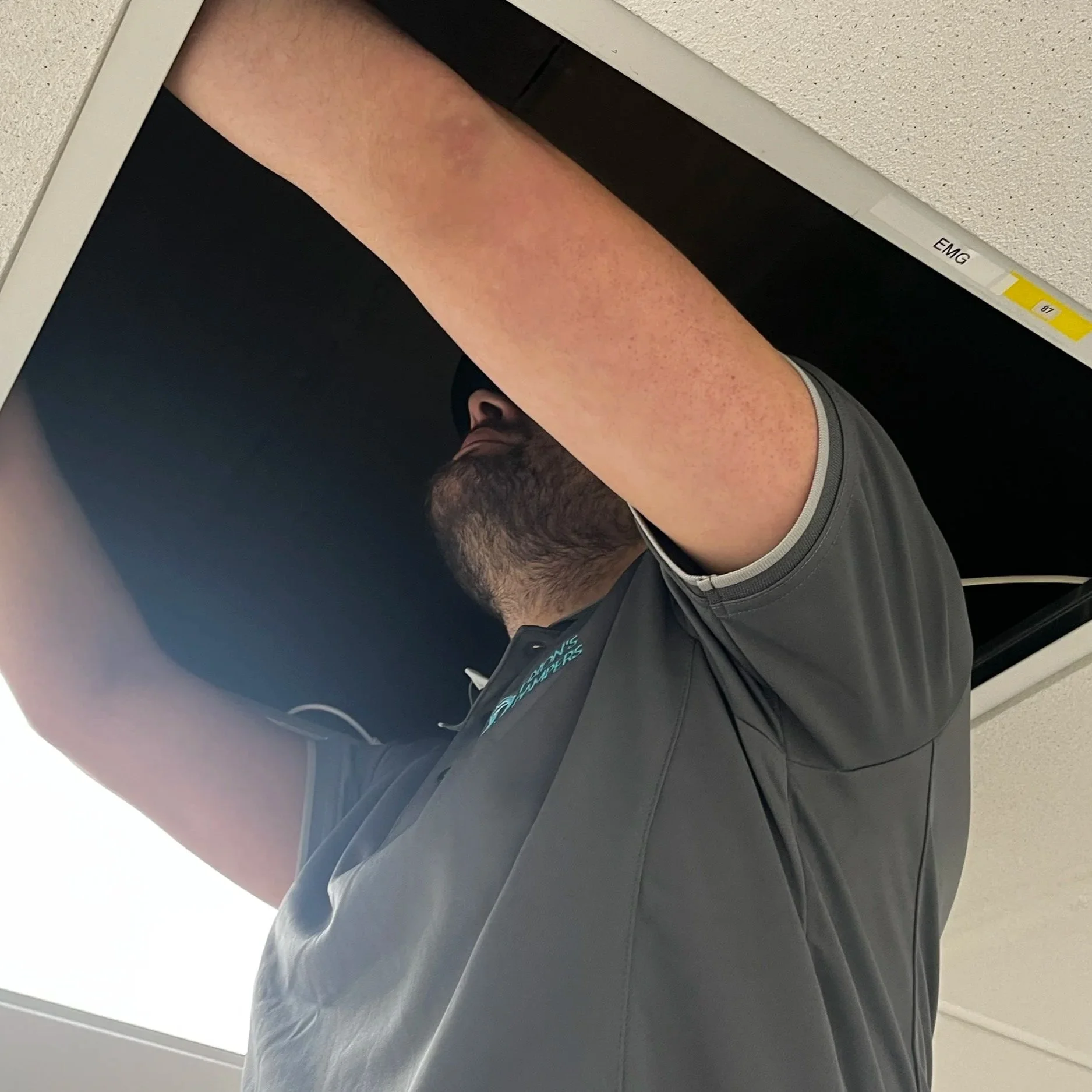 A man with a beard and wearing a gray Nike jacket is standing behind an open ceiling tile, reaching up and looking into the ceiling space.