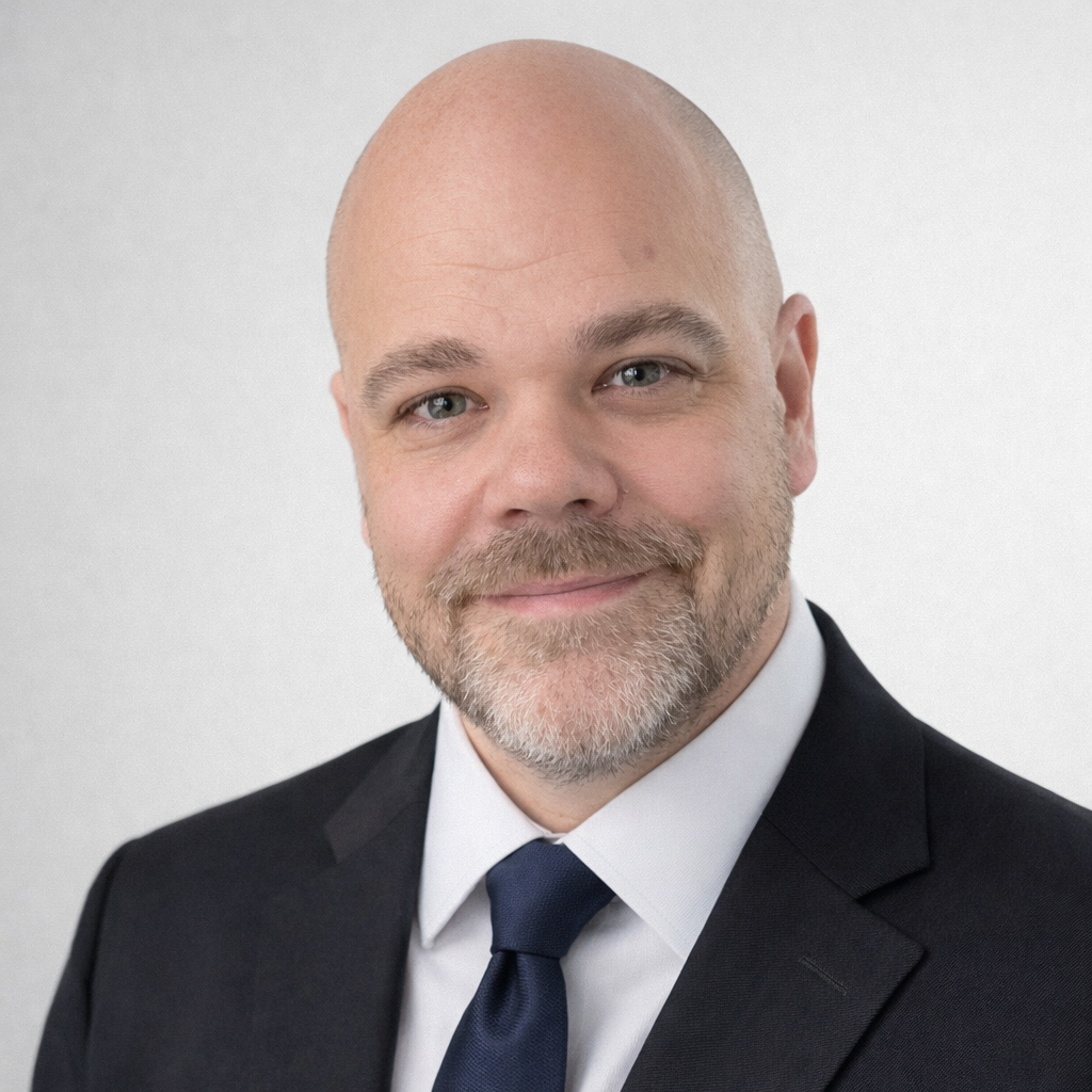 A professional headshot of a bald man with a beard, wearing a dark suit, white shirt, and blue tie, smiling against a plain white background.