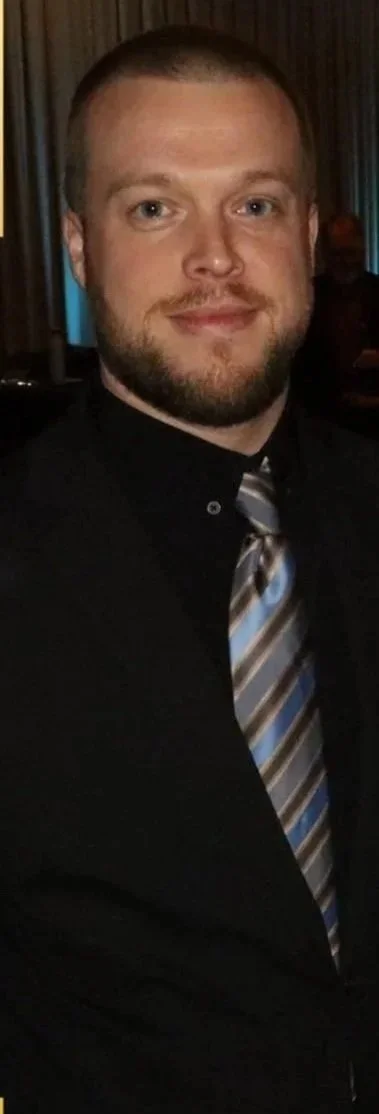 A man with a beard and short hair wearing a black shirt and a striped tie, smiling for the camera in a dimly lit indoor setting.