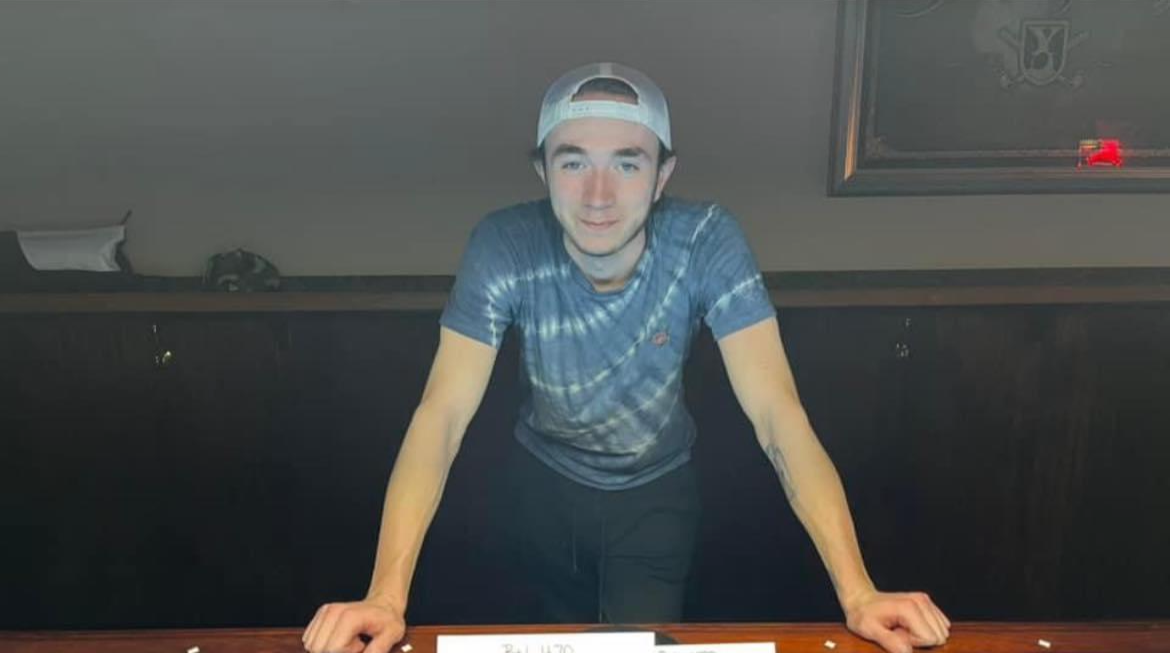 A young man wearing a striped blue t-shirt and a backward baseball cap leaning on a table with both hands, smiling at the camera in a dimly lit room.