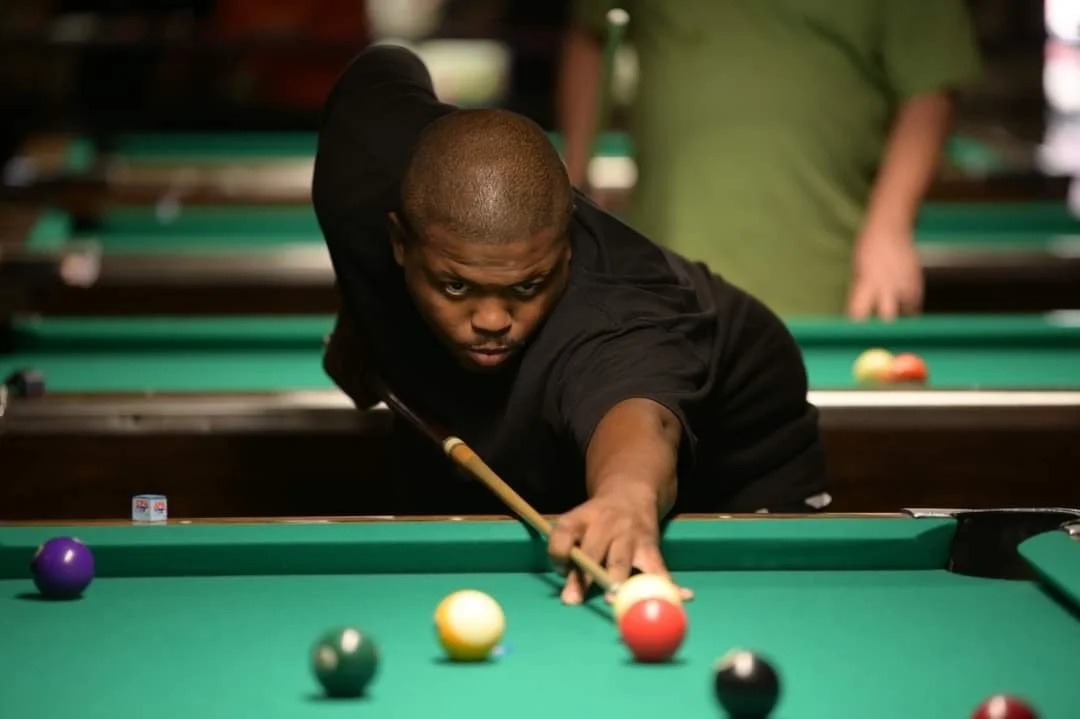 A man playing pool, aiming with a cue stick at a striped ball on a green pool table.