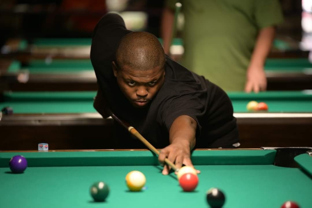 Young man in black shirt concentrating while playing pool. He is aiming to hit the red ball on a green pool table, with other balls scattered around.