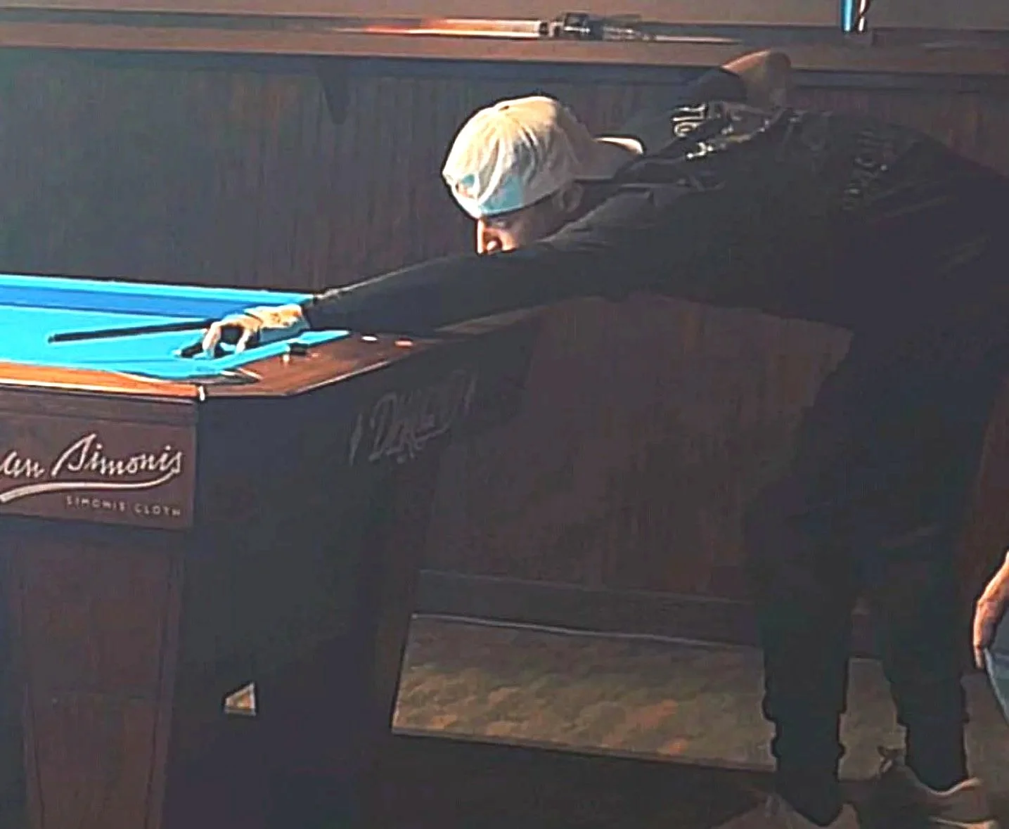 A person playing pool at a table with a blue felt surface, wearing a light-colored cap and black clothing, leaning over to take a shot.