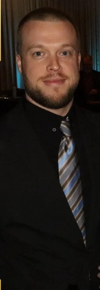 A man with short hair and a beard, wearing a black shirt and a striped tie, smiling at the camera in a dimly lit setting.