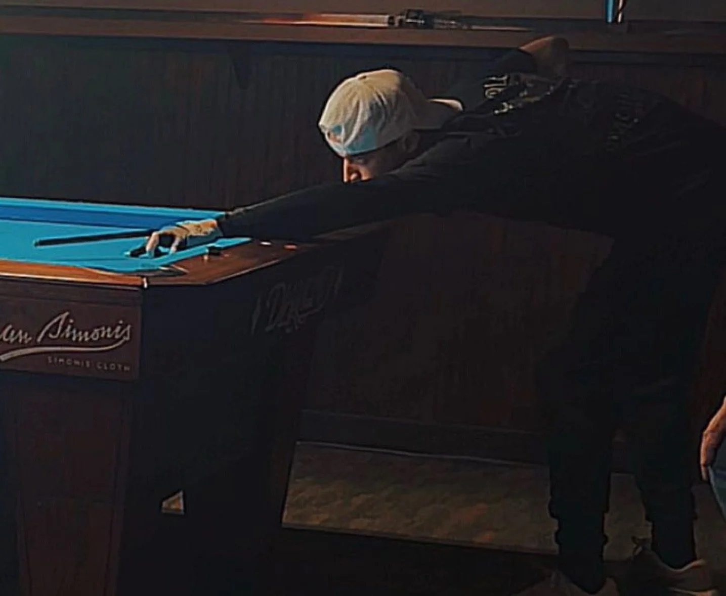 A person with blonde hair wearing a white cap, black jacket, and black pants, leaning over a pool table, aiming to hit a cue ball with a cue stick. The table has a blue felt surface and wood trim, with a sign reading ''Lou Simon's'' on the side.