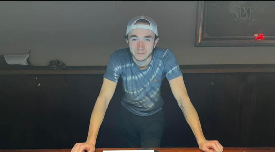 A young man wearing a backwards cap and a blue tie-dye shirt, leaning forward with hands on a table in a room with a dark cabinet and a framed picture on the wall.