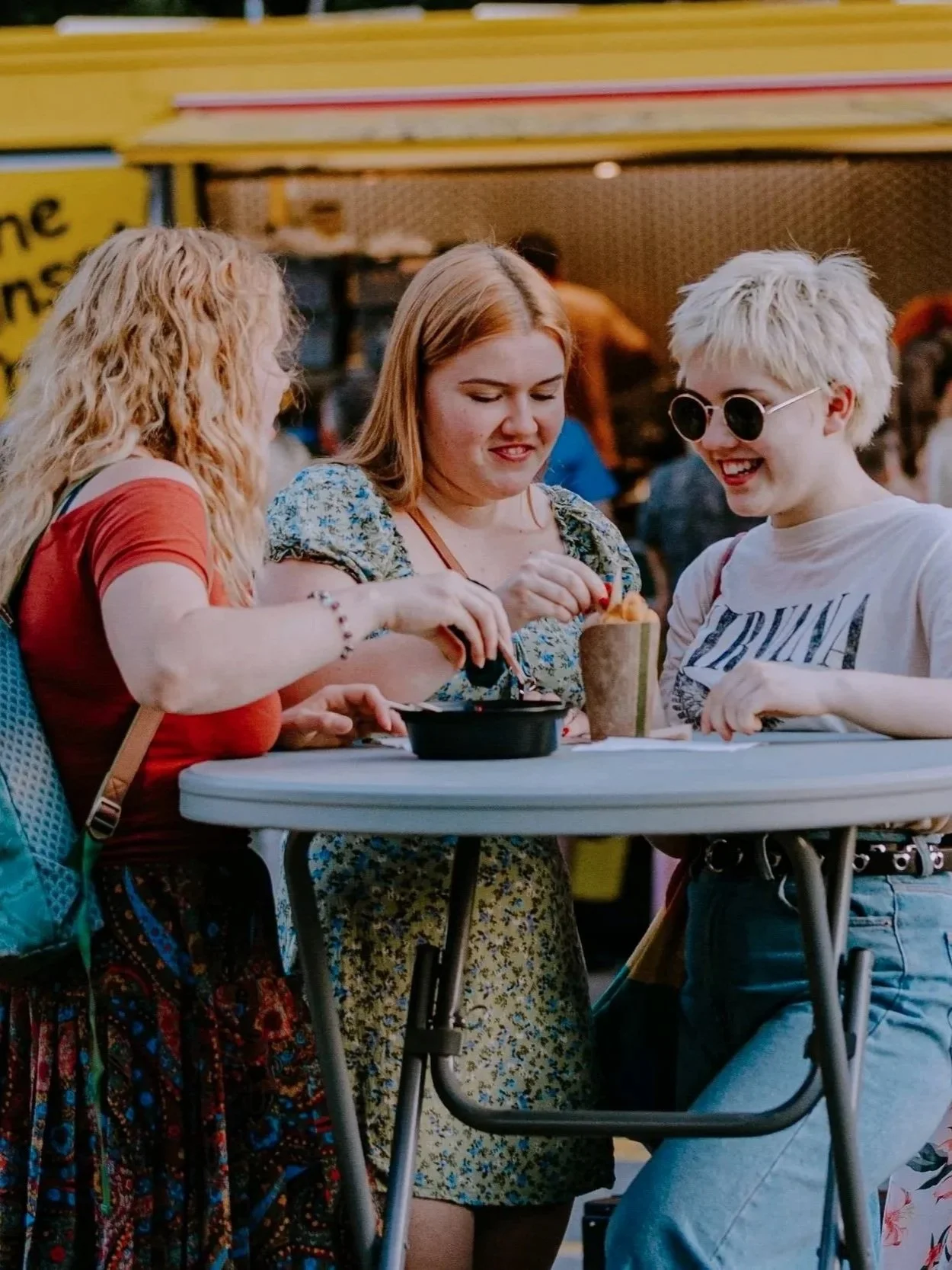 Three young women smiling and sharing a laugh at a table during an outdoor event, with food and drinks.