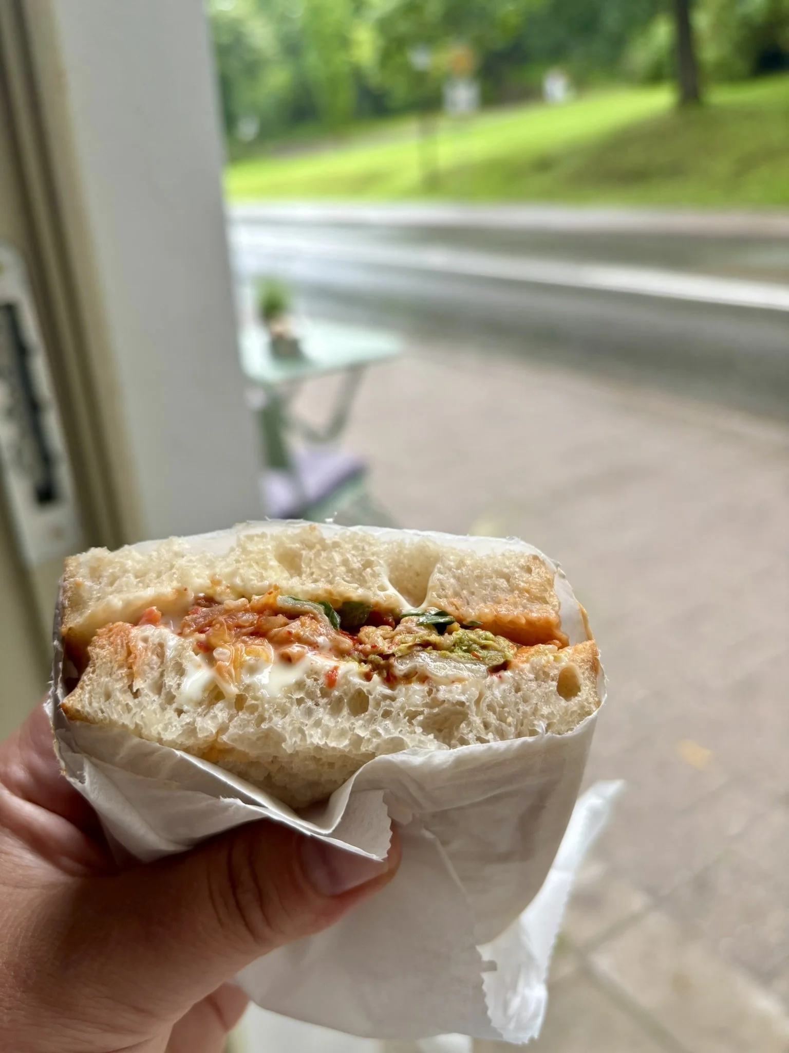 Close-up of a half-eaten sandwich with meat, vegetables, and sauce, being held in a person's hand near a window, with a blurred outdoor street and greenery in the background.