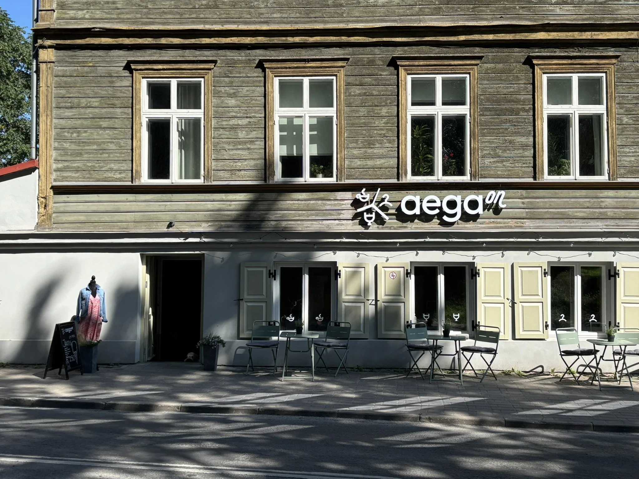 A building with a two-story wooden exterior and multiple windows on the upper floor. The ground floor has open shutters, outdoor seating, and a sign that reads 'Aega on' with a clock logo.