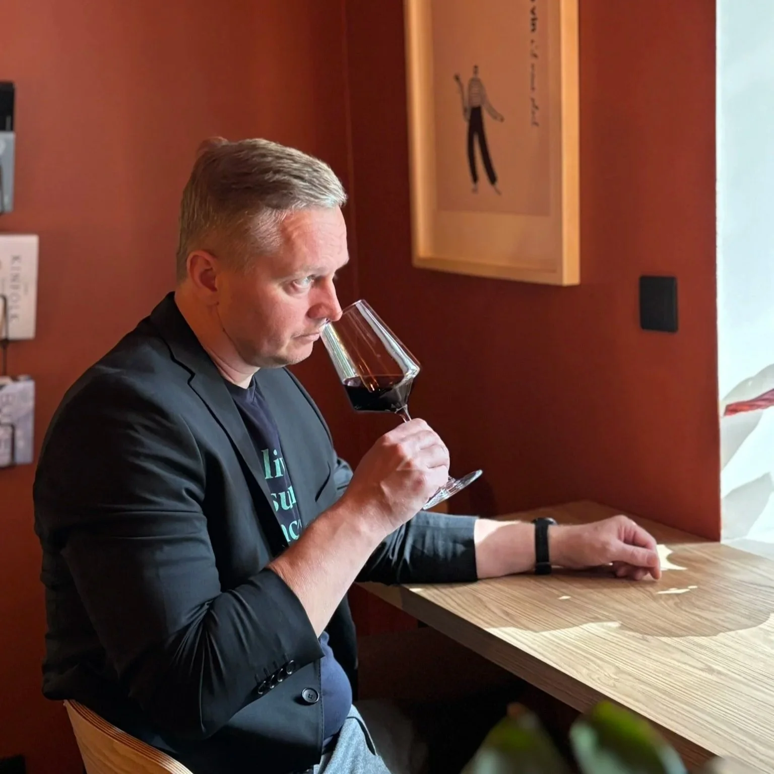 A man in a black blazer and dark t-shirt is sitting at a wooden counter, holding a glass of red wine close to his nose and smelling it.