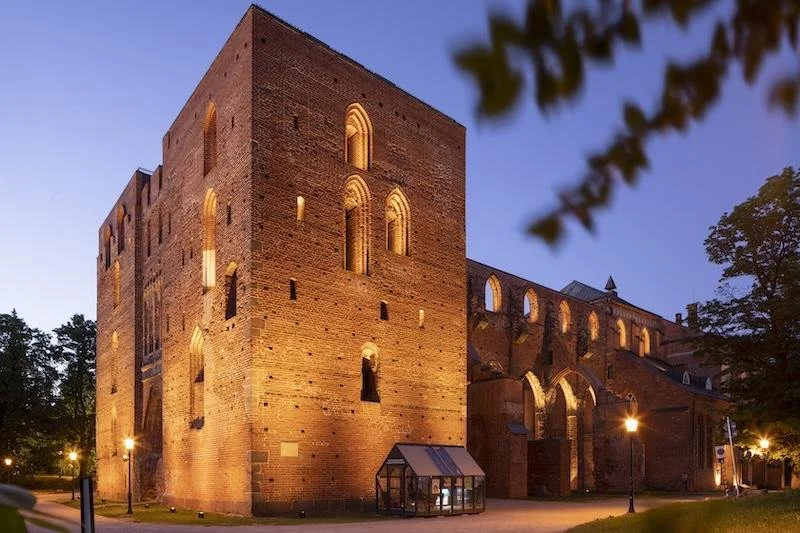 A historic brick castle or fortress during twilight, illuminated by exterior lights, with trees and a small glass structure in the foreground.