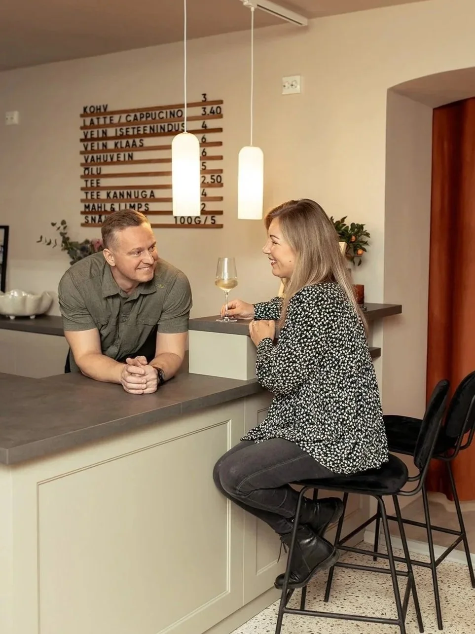 A man and woman in a cafe, engaging in conversation, with the woman holding a glass of wine. The cafe has a menu on the wall and modern pendant lighting.