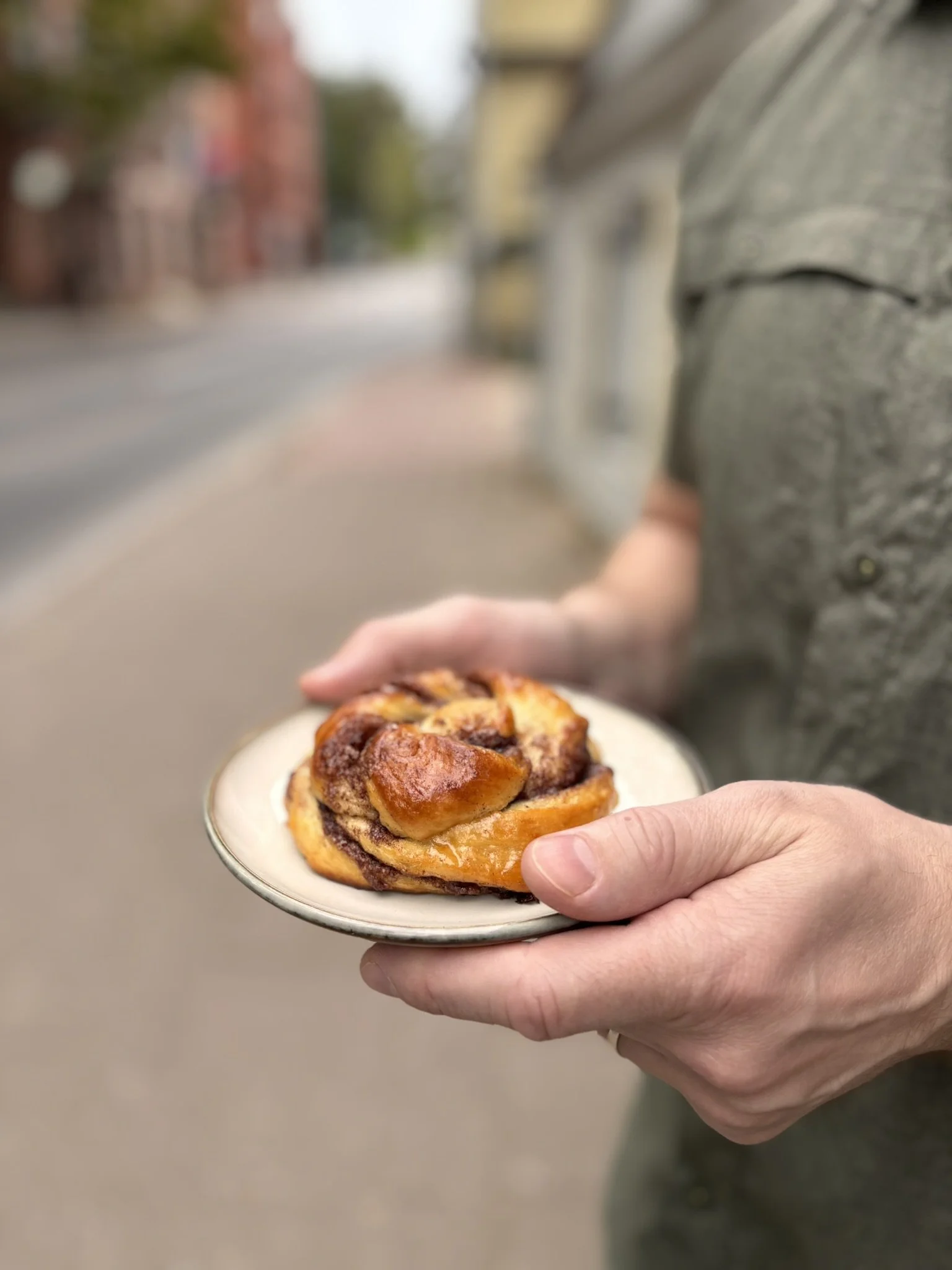 Person holding a small plate with a cinnamon roll outdoors on a sidewalk.