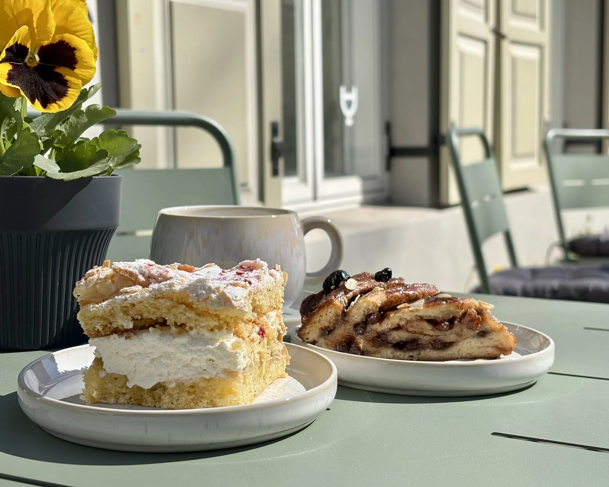 Slices of layered cake and a slice of apple cinnamon pastry on white plates, with a white mug and potted pansy flower on a green outdoor table, in a patio setting.