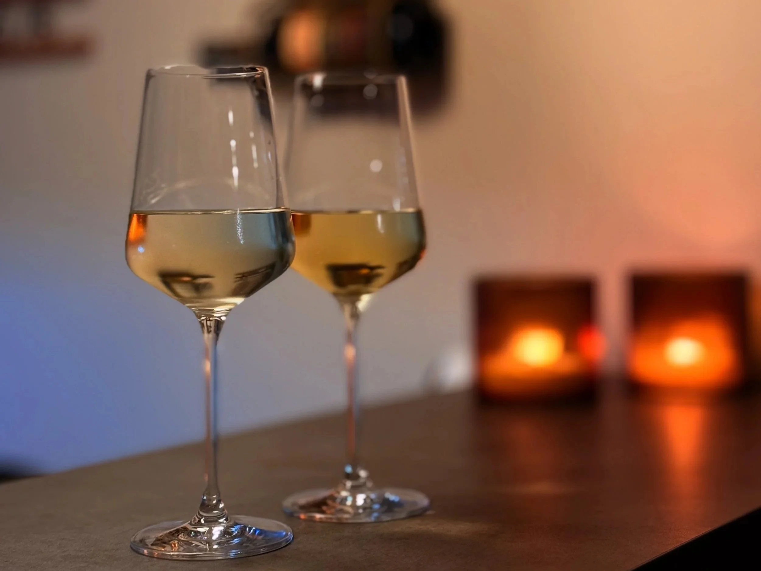 Two glasses of white wine on a table with blurred candles in the background.