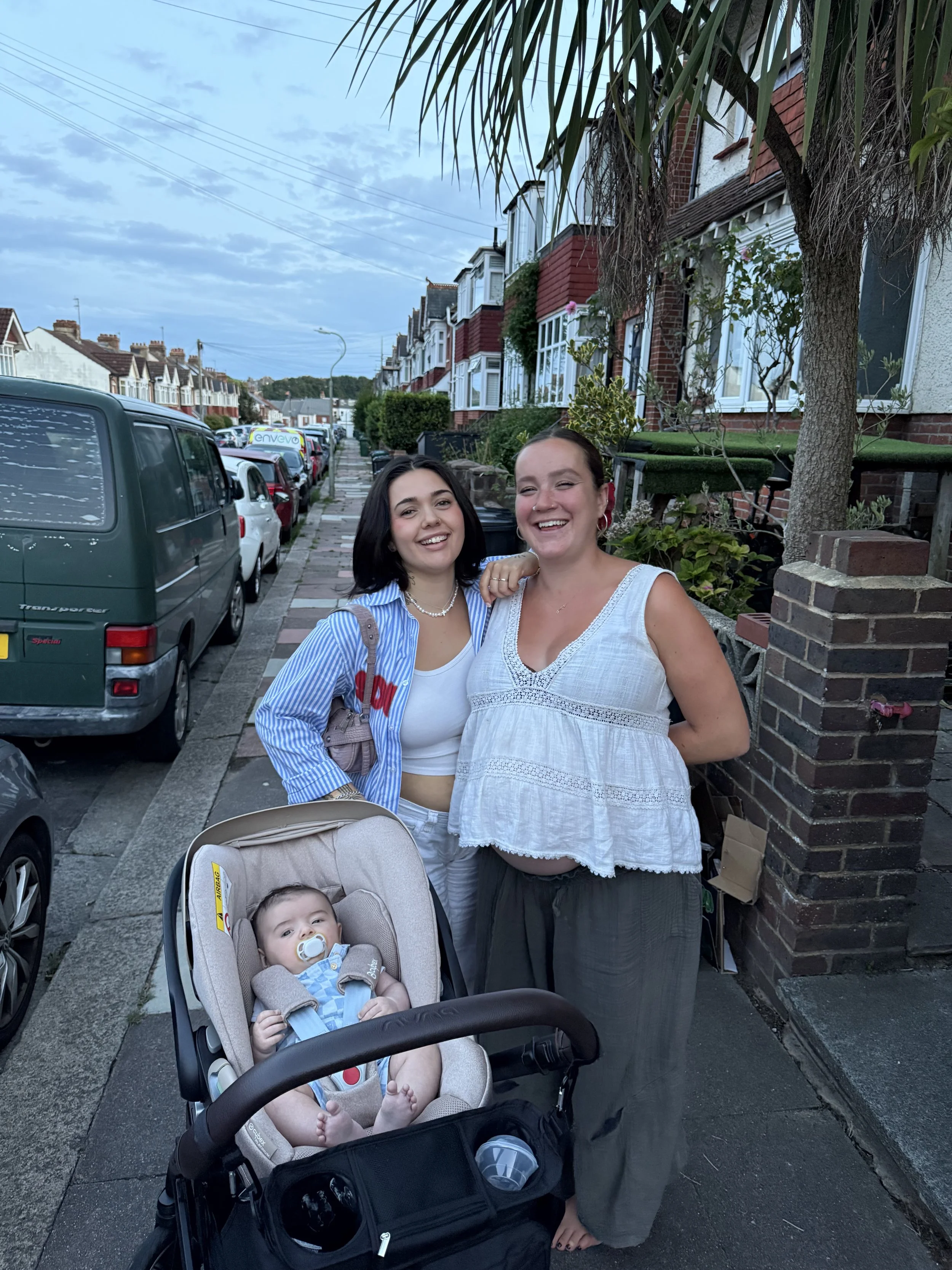 Two women smiling with one arm around each other, standing on a residential sidewalk, with a baby in a stroller in front of them, under a cloudy sky.