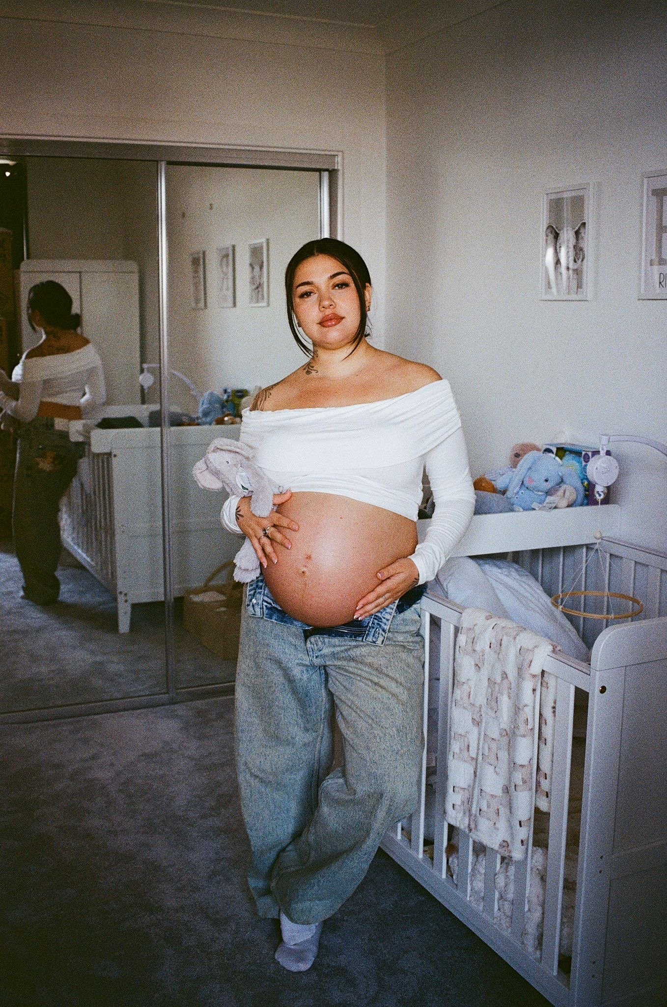 Pregnant woman standing in a nursery holding her belly, wearing jeans and a white off-shoulder top, with a woman in the background near a crib and a mirror.