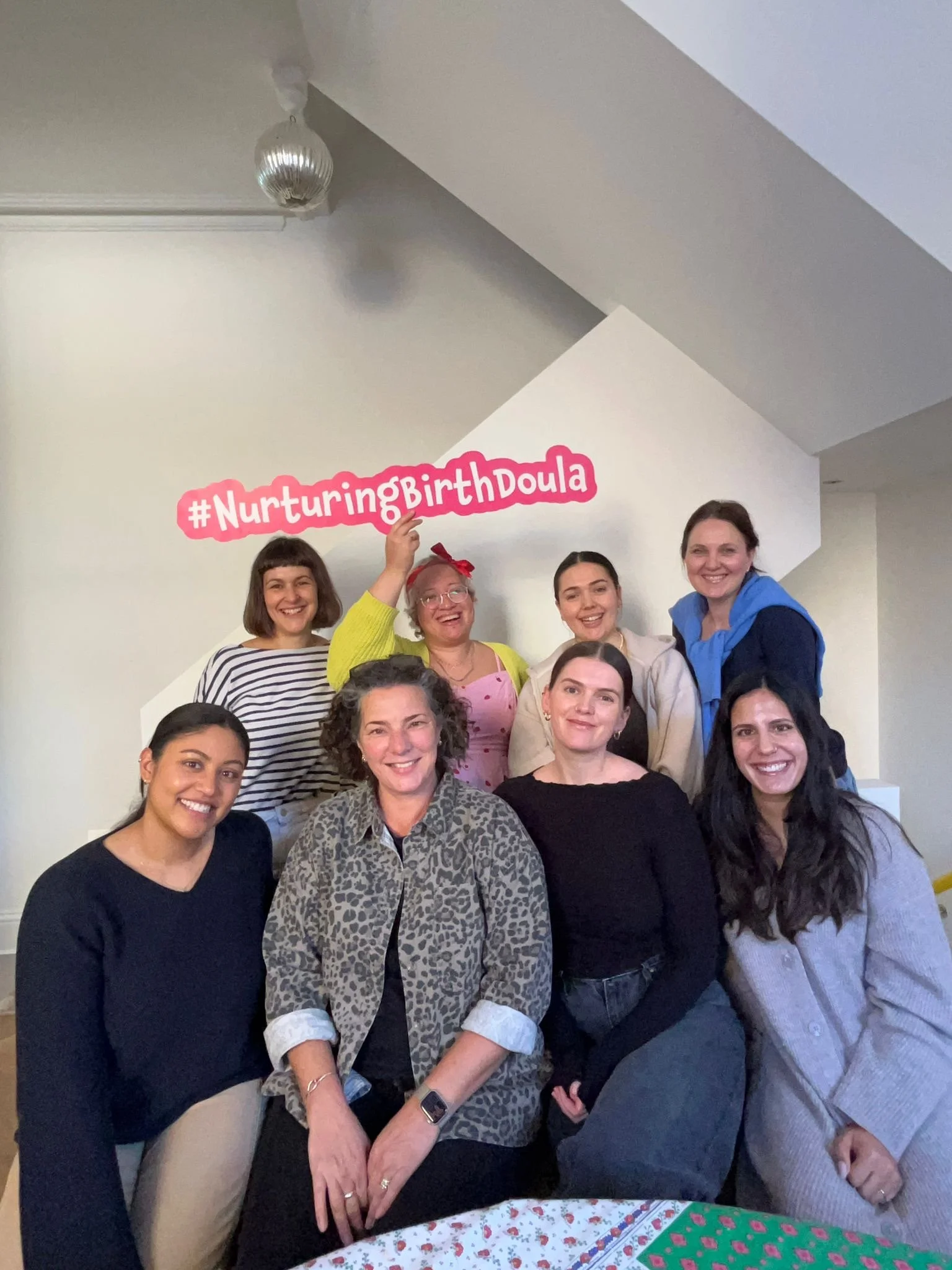 Group of eight women smiling at a gathering, with one holding a pink sign that says #NurturingBirthDoula, in a room with white walls and a staircase in the background.