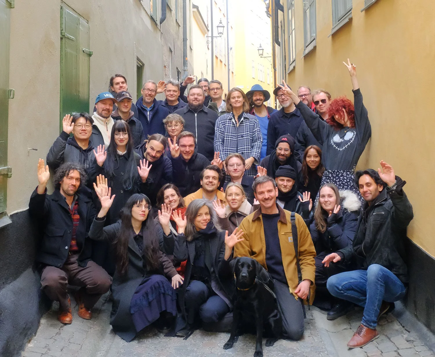 Group of people posing on a narrow street, some waving, with a black dog sitting in front. The group includes men and women of various ages, wearing casual jackets and sweaters. The street is lined with colorful old buildings.
