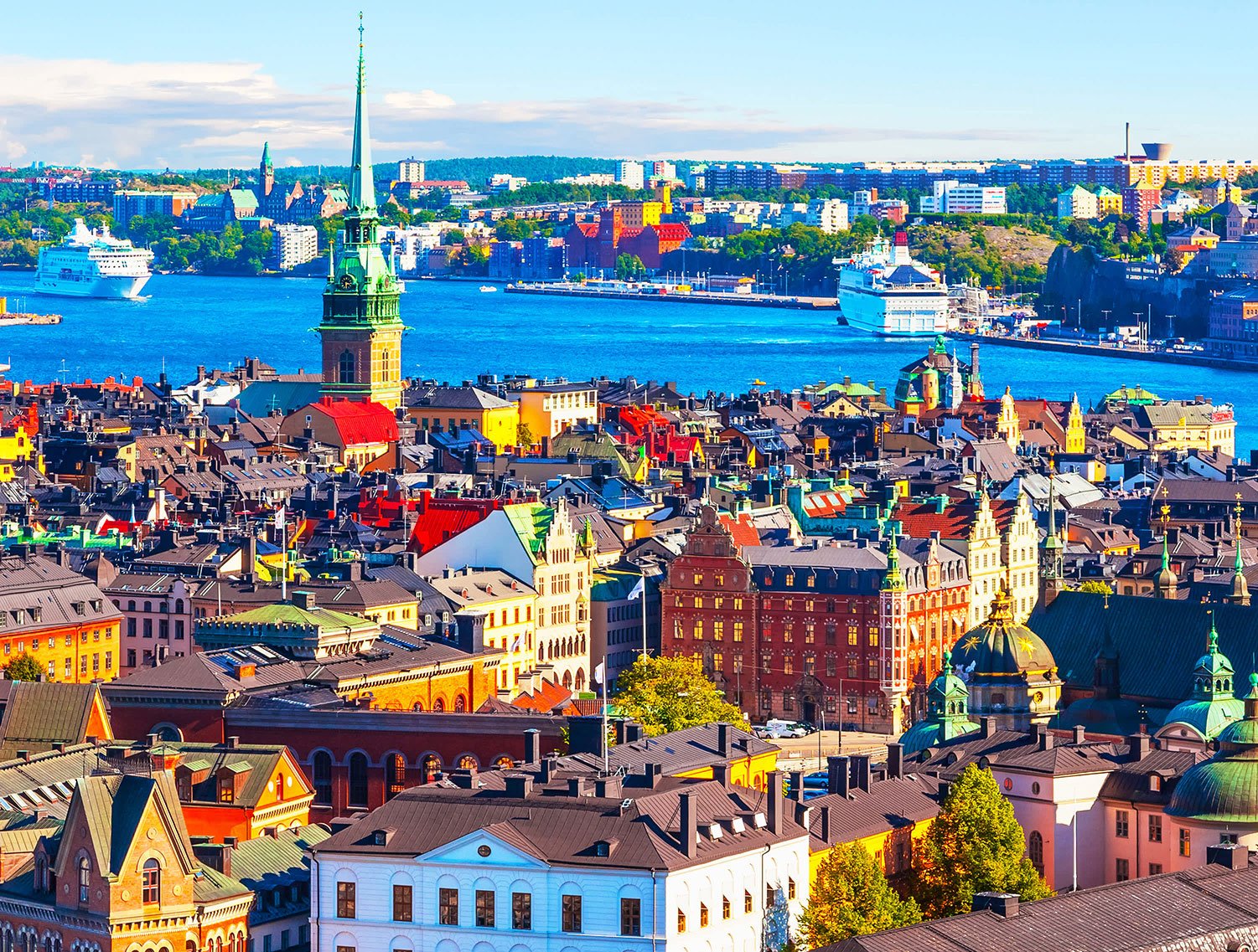 Aerial view of Stockholm, Sweden, with colorful historic buildings, a large body of water, and ships, including a cruise ship and ferries, under a partly cloudy sky.