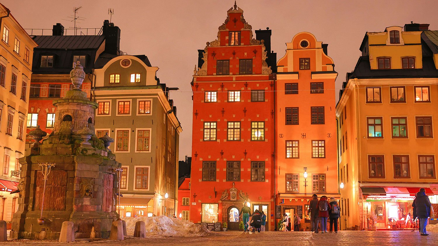 Nighttime scene of colorful historic buildings in a European city square with people walking, a stone fountain in the foreground, and warm lighting illuminating the buildings' facades.